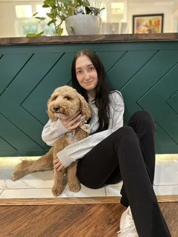 Young woman sitting on the floor, hugging a small, fluffy, tan-colored dog in front of a teal-colored wall with plants and artwork in the background.