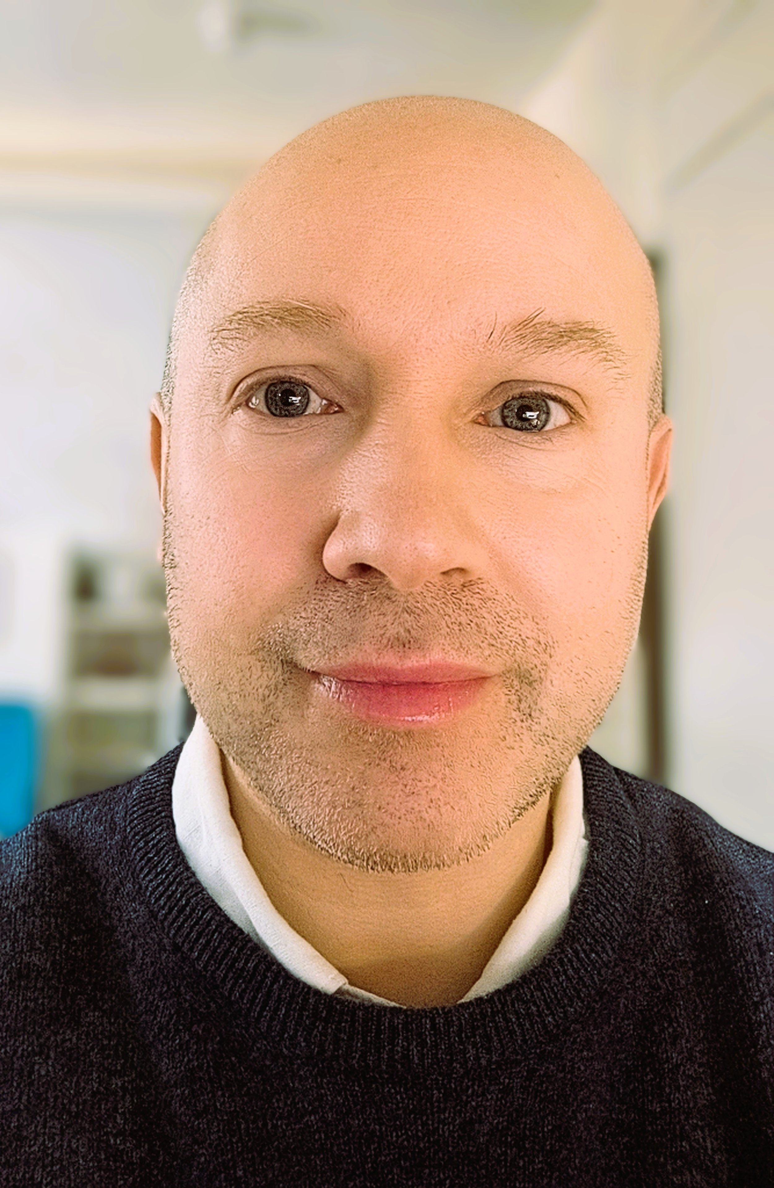 A close-up photo of a man with a bald head, light skin, and blue eyes, wearing a dark sweater over a collared shirt.