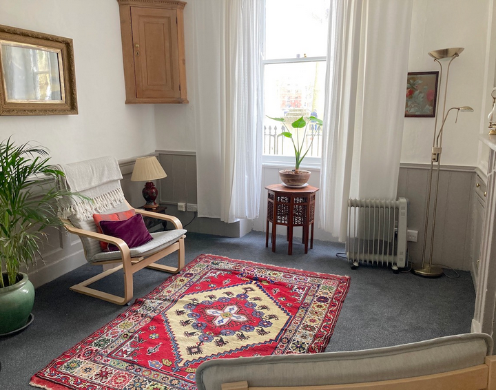 A cozy living room with a gray carpet, a red and beige patterned rug, a wooden armchair with a white cushion and purple pillows, a side table with a lamp, a large window with sheer white curtains, a potted plant on a small table, a standing lamp, and