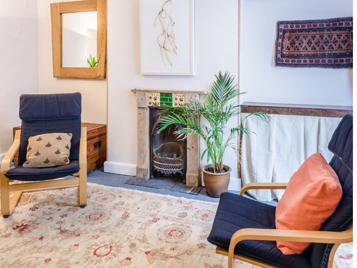 Cozy living room with two navy armchairs, a fireplace with green tiles and wooden mantel, a potted palm, colorful rugs, and wall art.