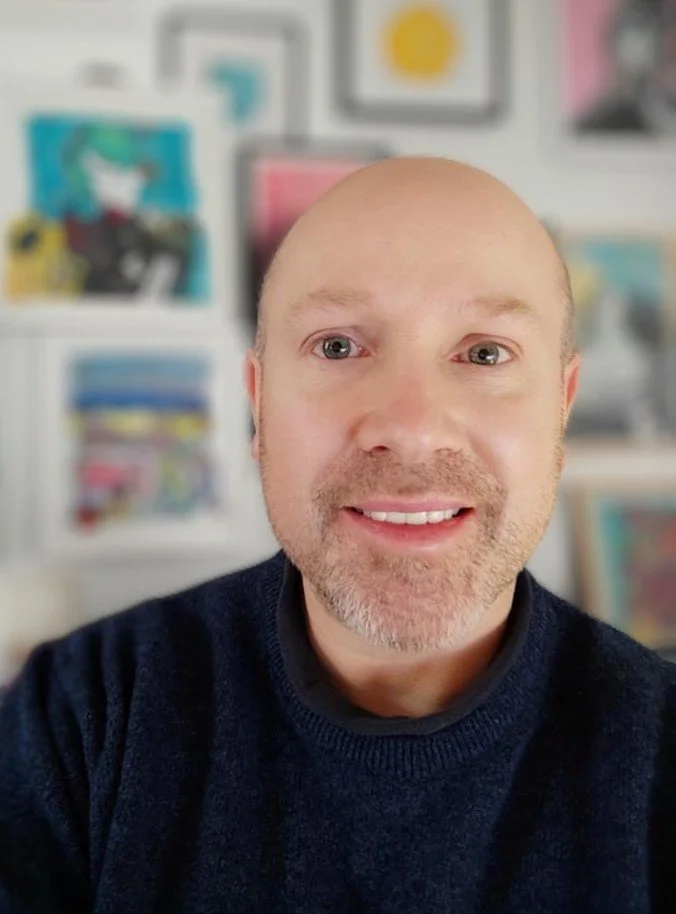 A man with a bald head and a beard taking a selfie in a room with colorful artwork on the wall behind him.