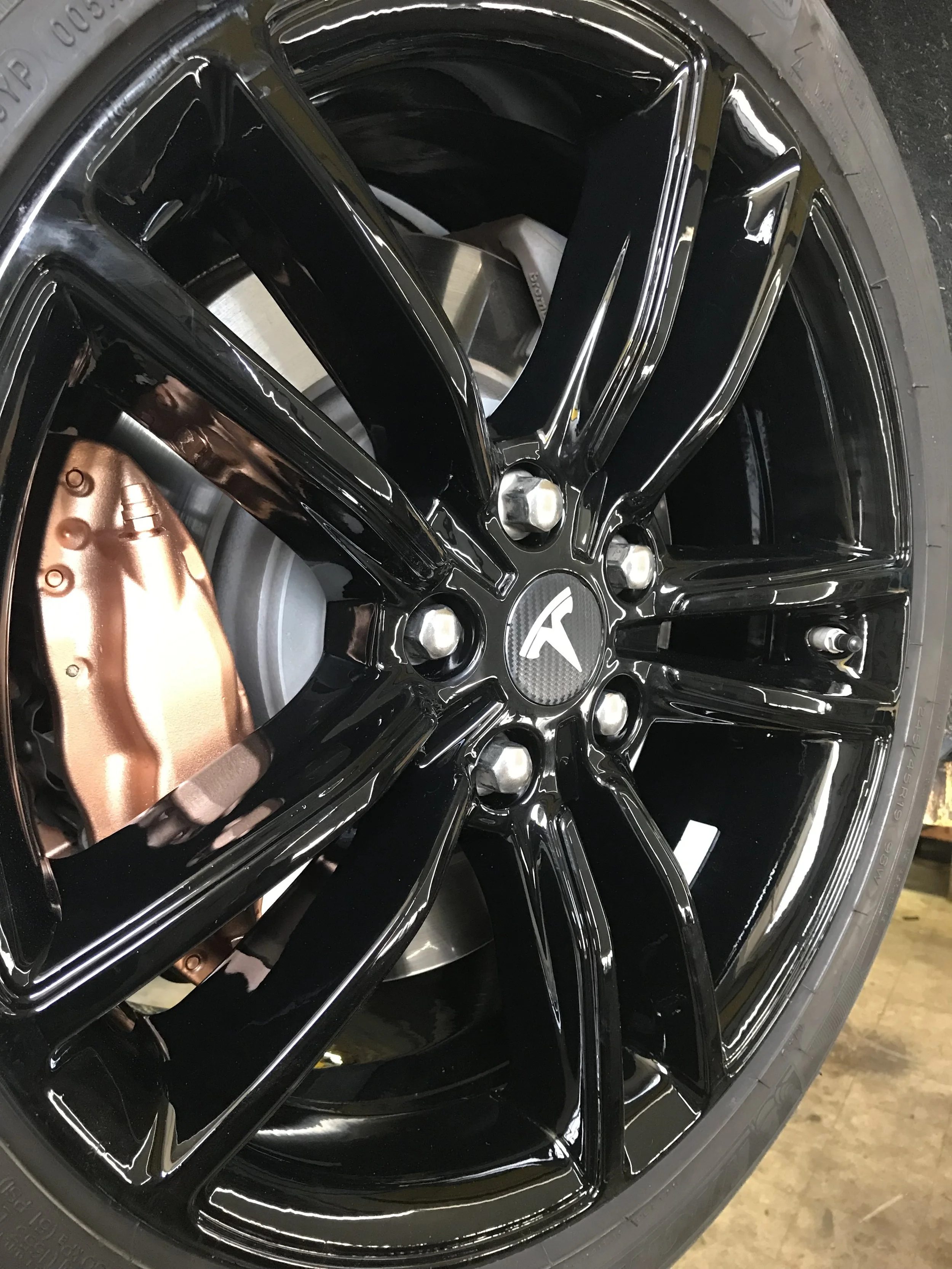 Close-up of a black Tesla alloy wheel with a copper brake caliper and a tire, showing the Tesla logo on the wheel center cap.