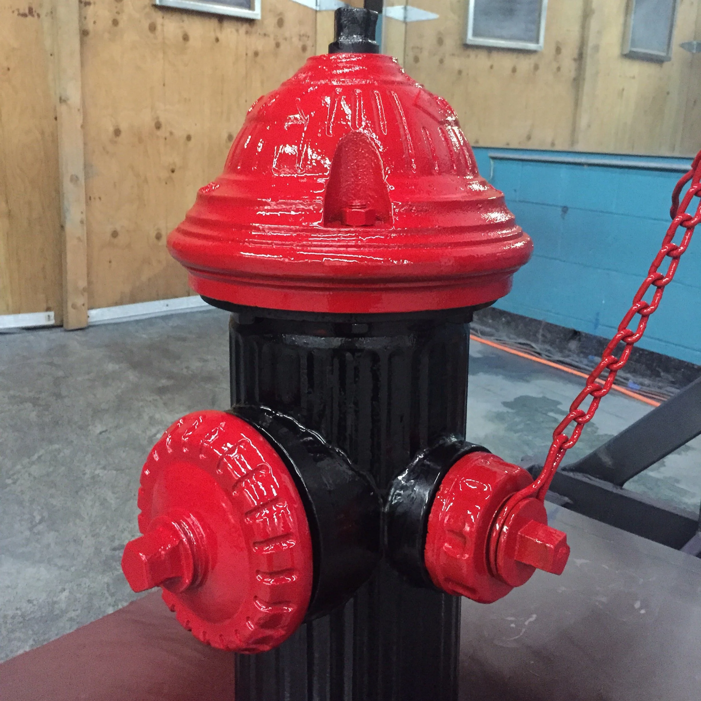 A fire hydrant painted with red and black paint, with the spray nozzles covered with red caps, located indoors with wooden and blue walls.
