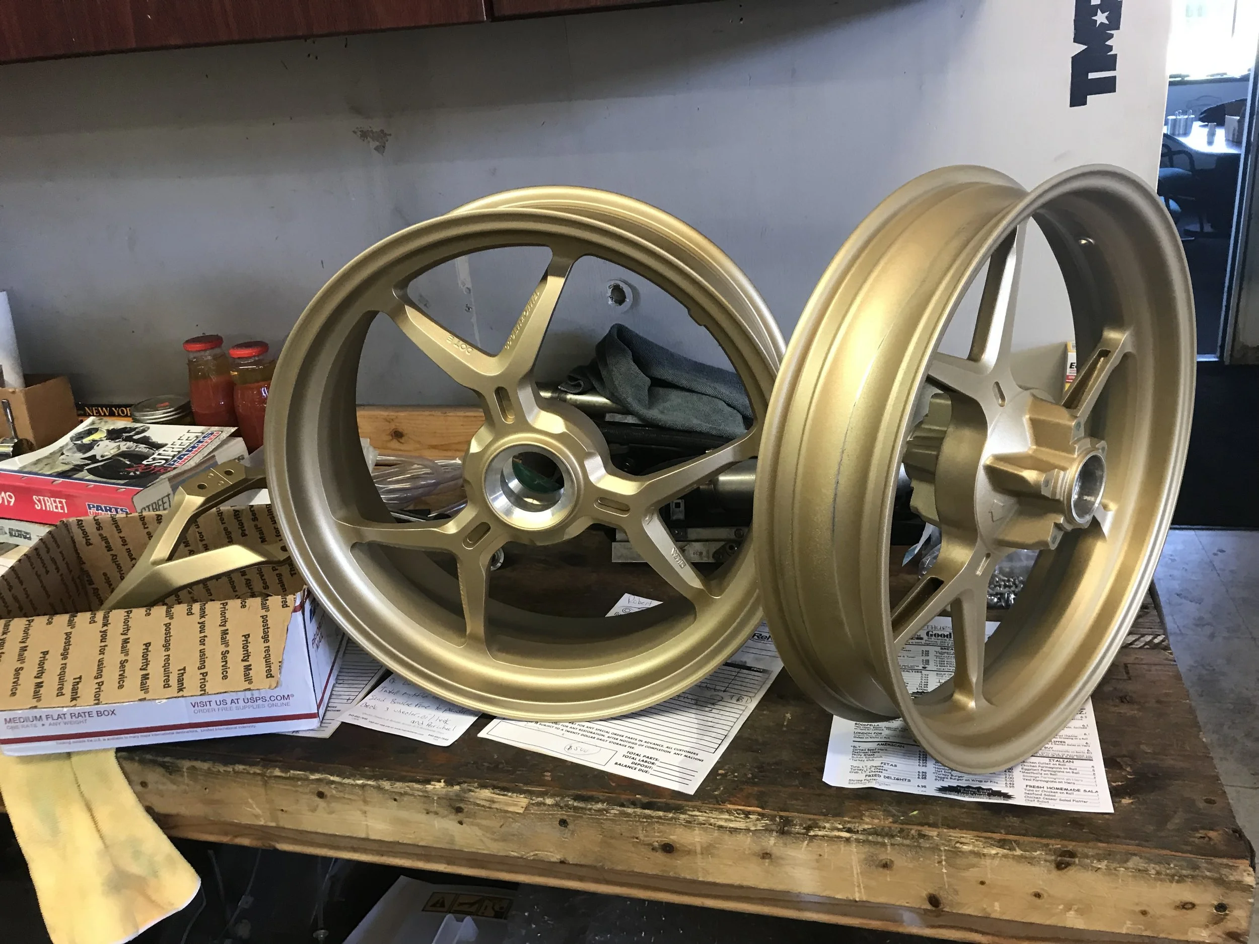 Two gold-colored motorcycle wheels on a cluttered workbench with papers, a box, and bottles in the background.