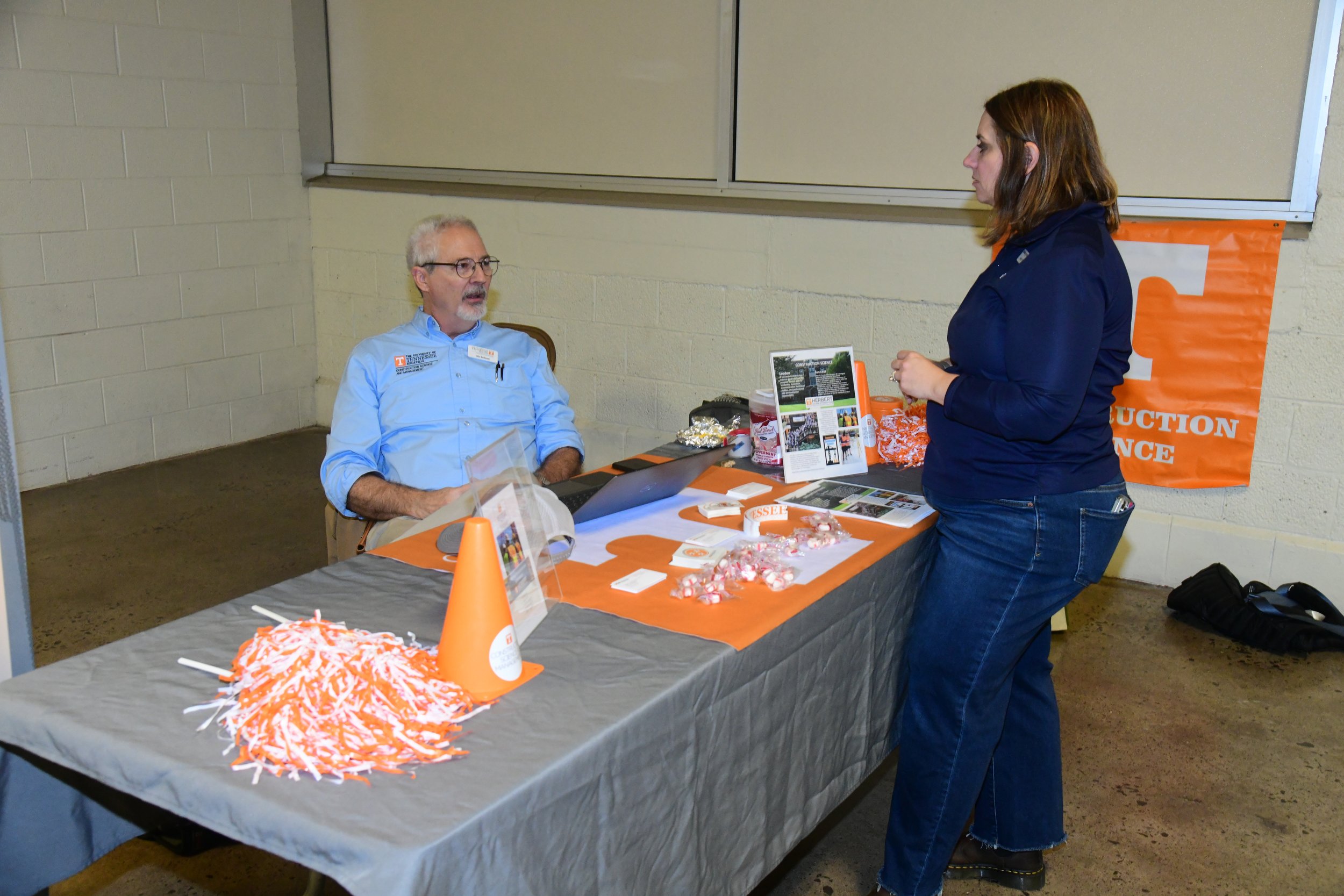 A man and a woman are having a conversation at a career fair booth decorated with orange and gray tablecloths, and promotional materials, in an indoor setting.