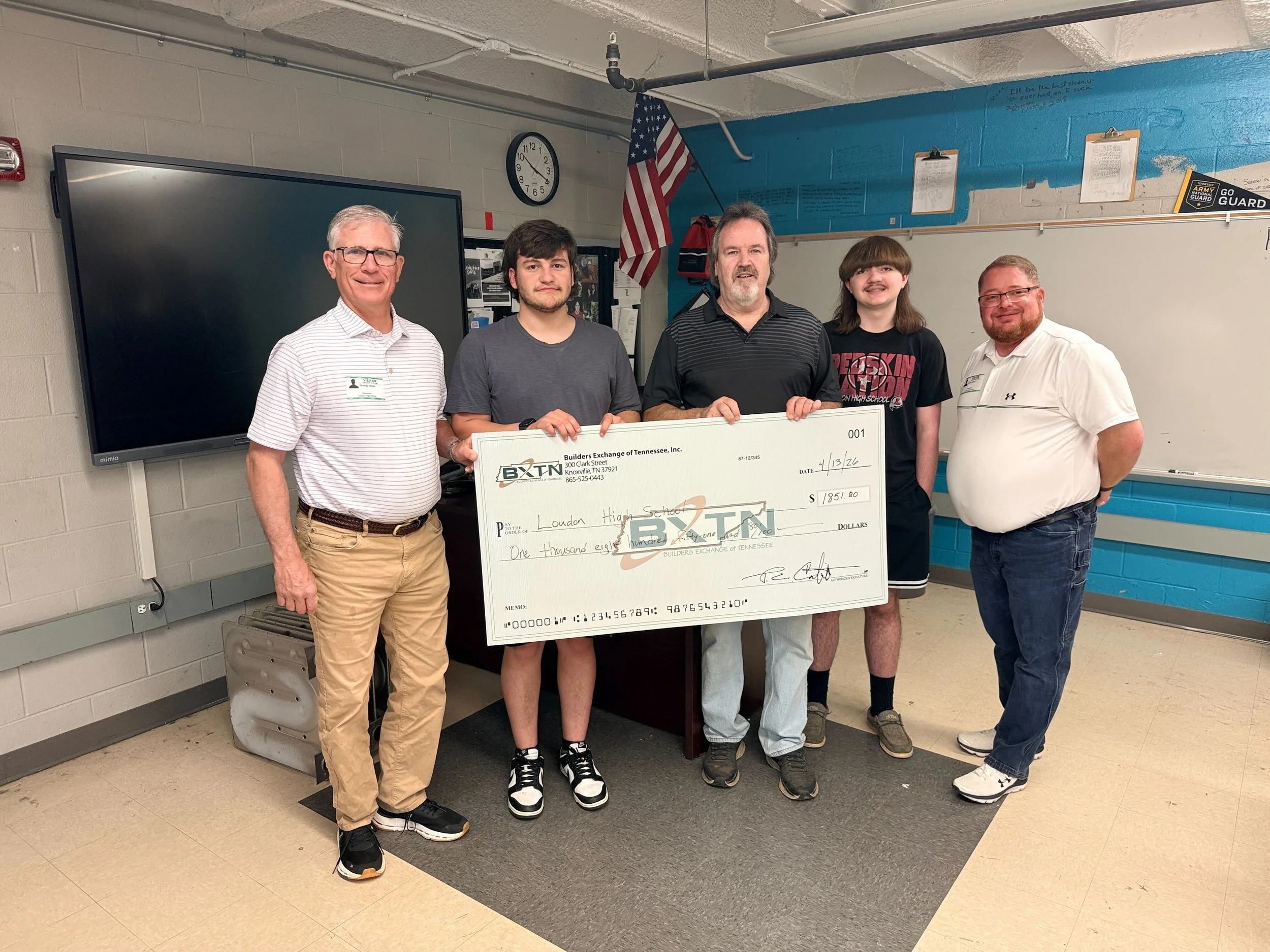 Group of five men standing in a classroom, holding a large ceremonial check. The classroom has a whiteboard, a large display screen, and an American flag. The check is from Builders Exchange of Tennessee and is made out to London High School for $1,851.80, dated April 13, 2026.