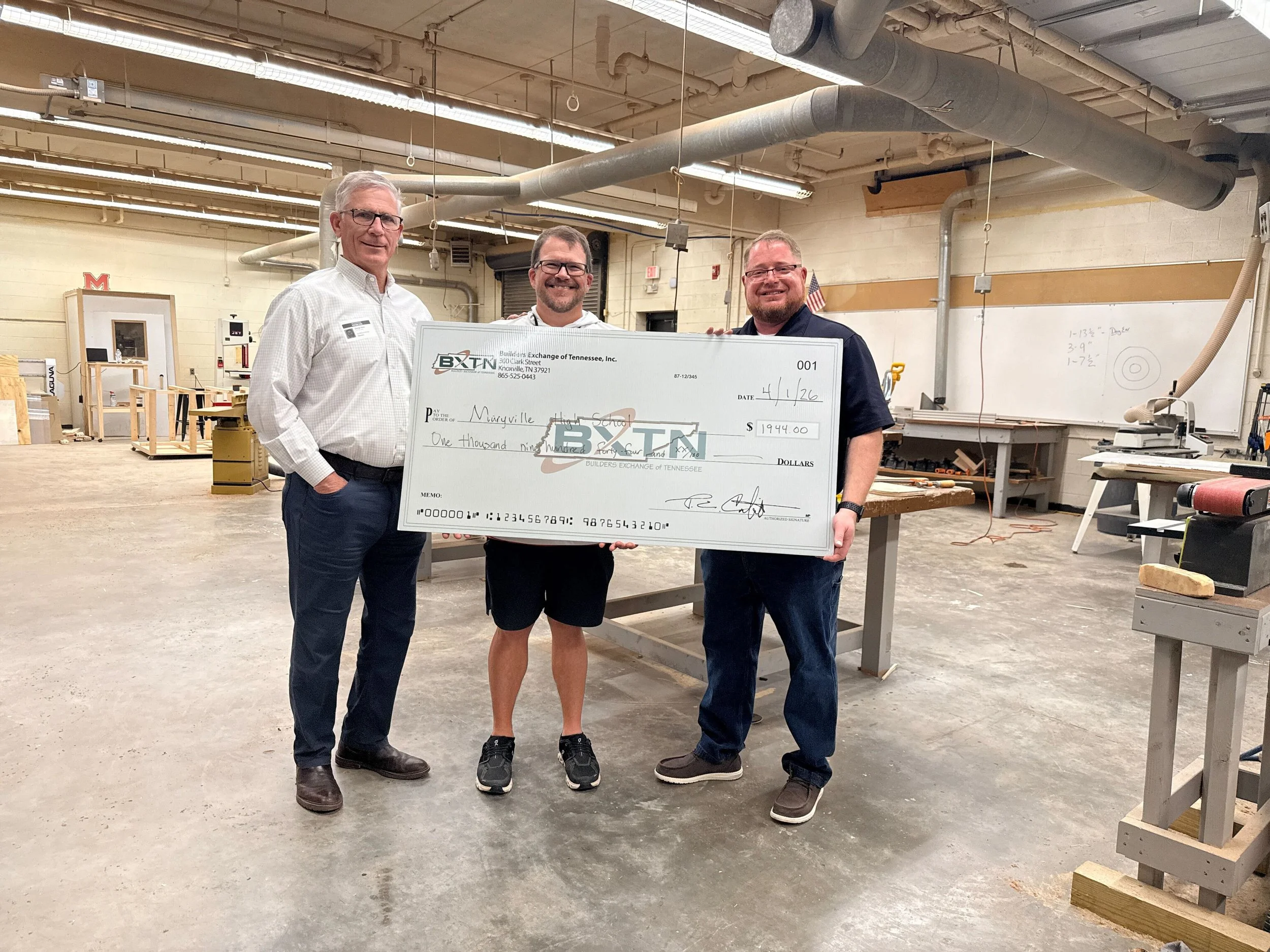 Three men standing in a workshop holding a large check. Two are wearing business attire and one casual clothing. The workshop has tools, whiteboard, and construction materials in the background.