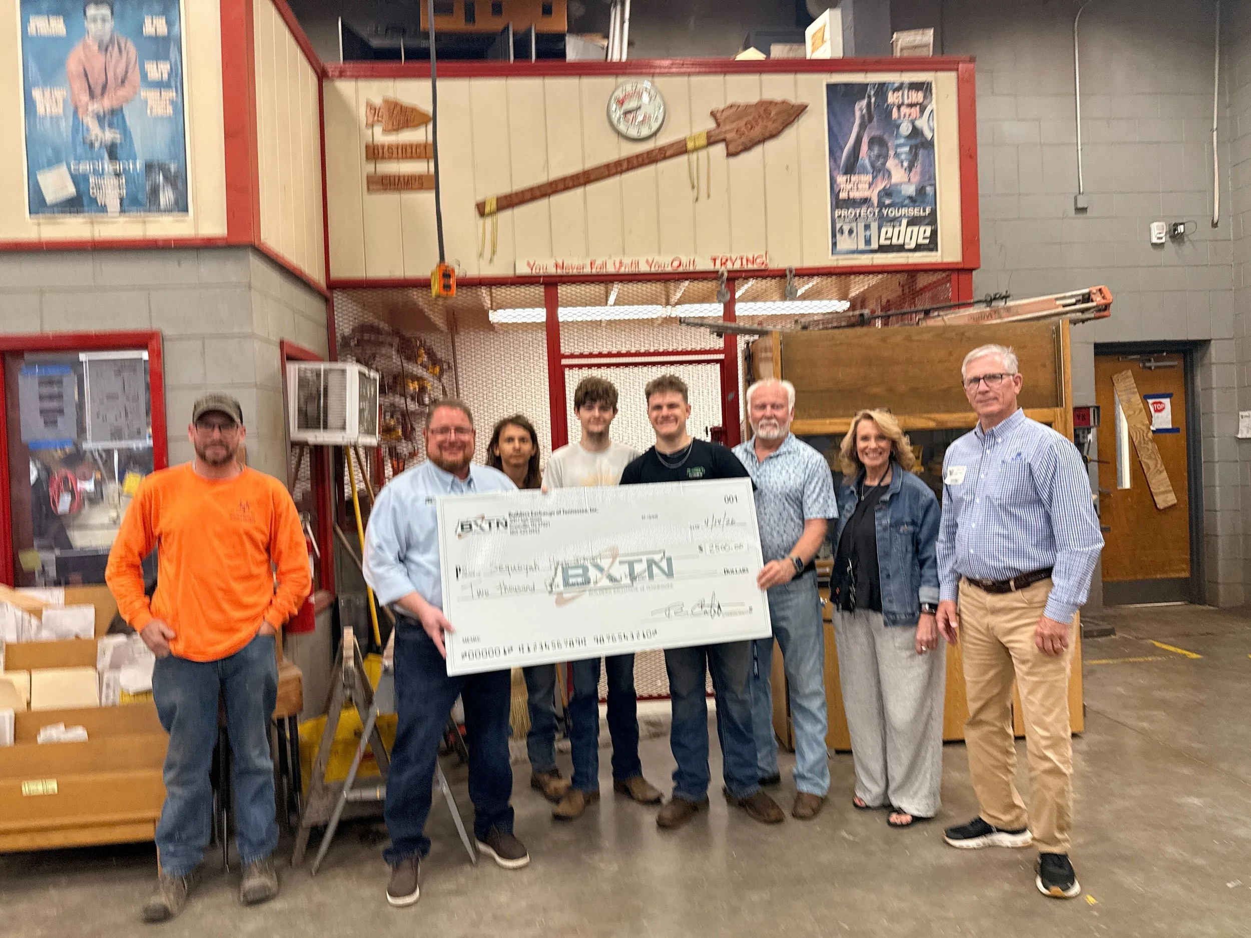 A group of seven people standing in a workshop holding a large check. Behind them are industrial shelves and tools, with posters and a clock on the wall.