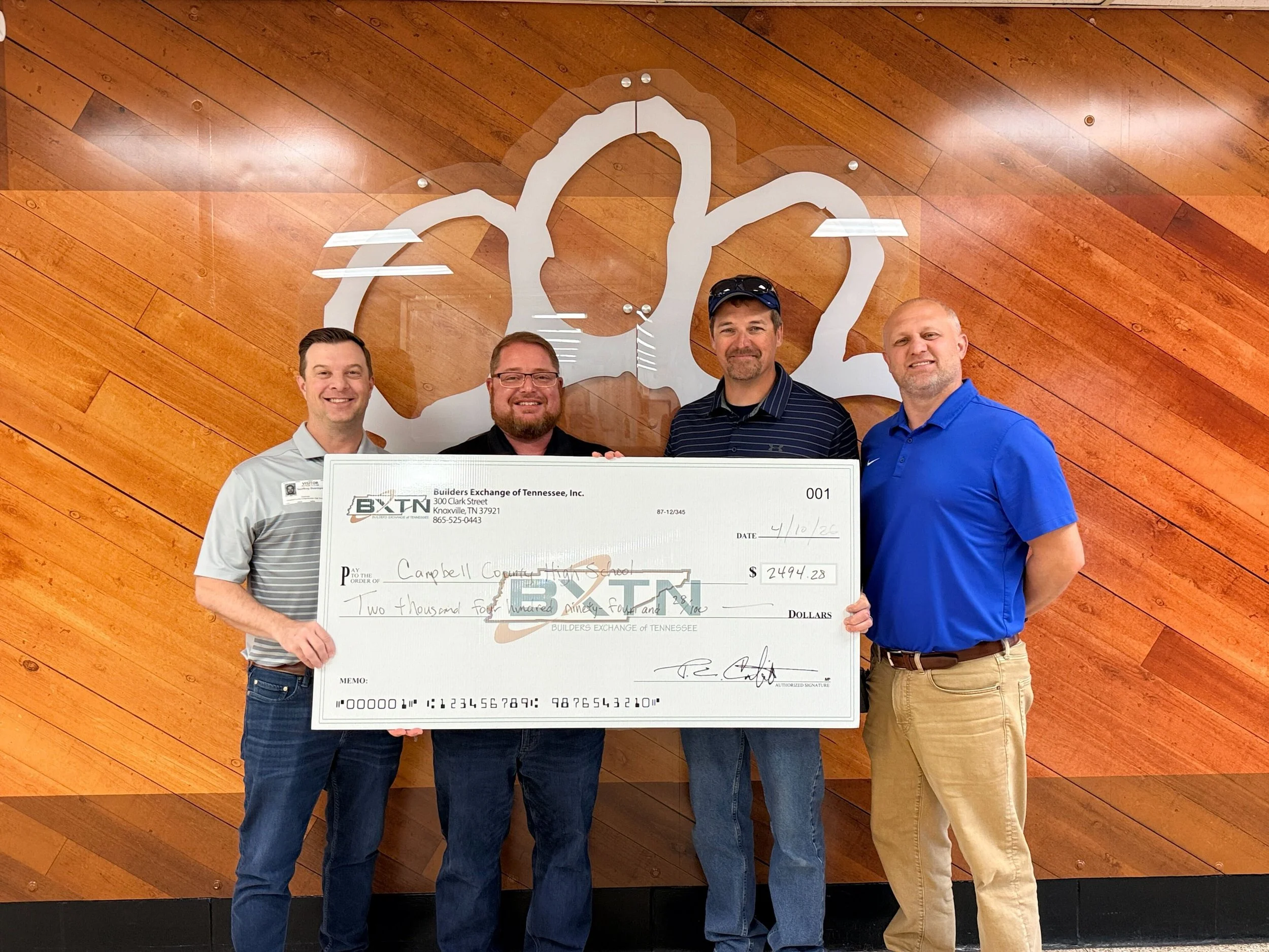 Four men standing in front of a wooden wall with a large white logo behind them. They are holding a giant check for $2,494.28, made out to Campbell County High School, from Builders Exchange of Tennessee.