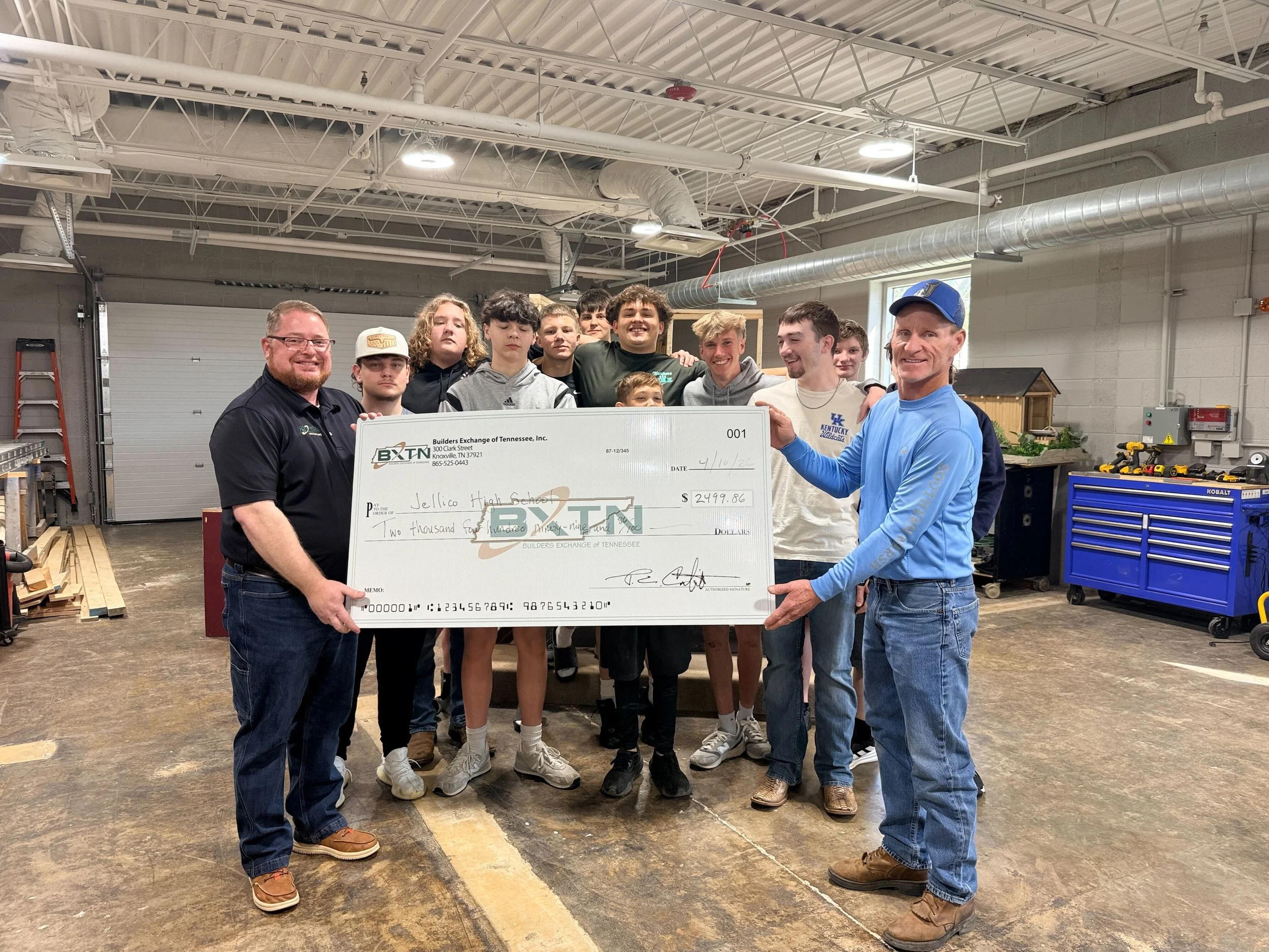 A group of young people and two adults standing inside a workshop holding a large check made out to Jellico High School for $2,499.86. The workshop has tools and workbenches, with a garage door and shelves in the background.