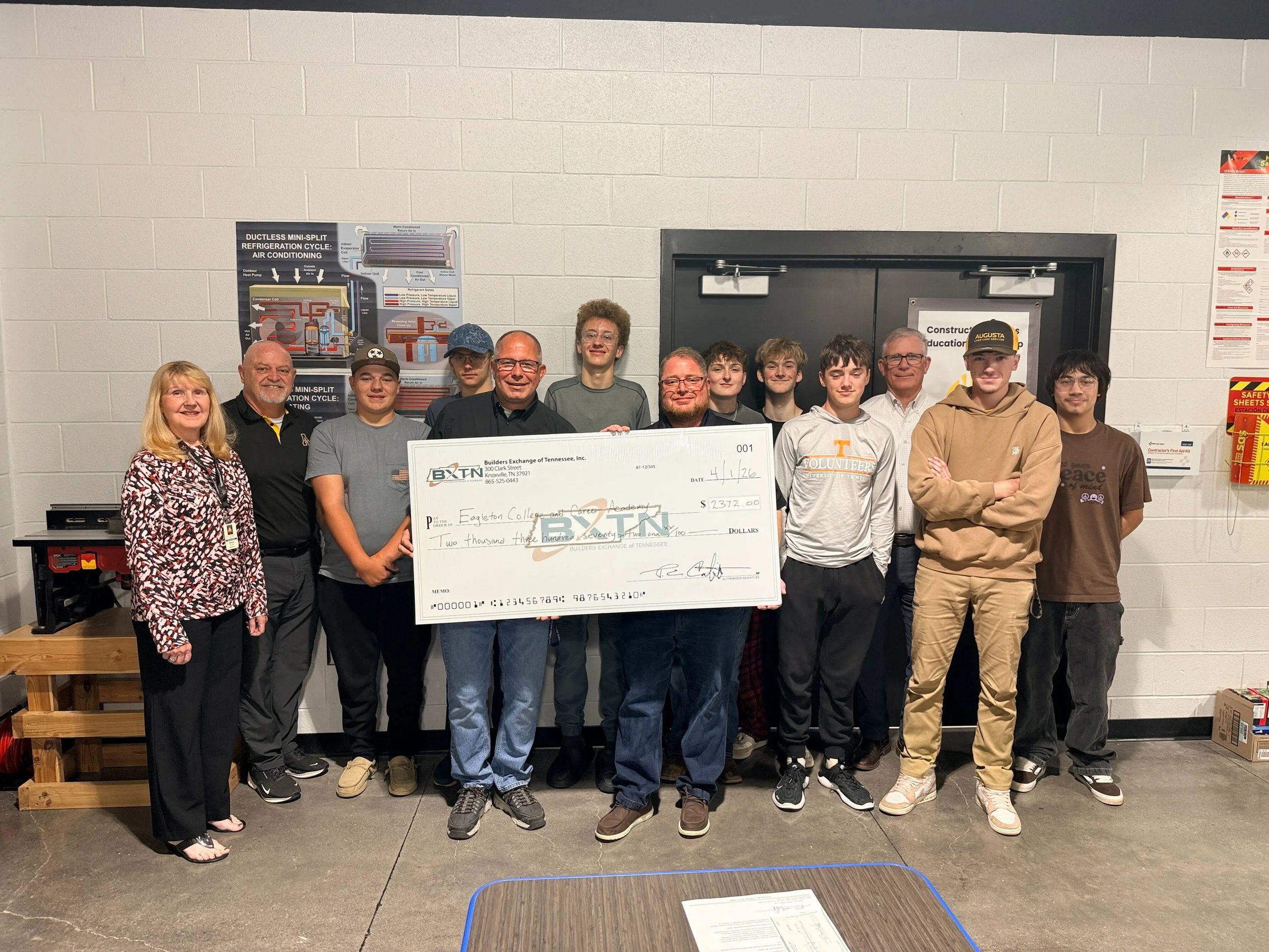 Group of people standing indoors, holding a large check, with a wall and posters behind them.