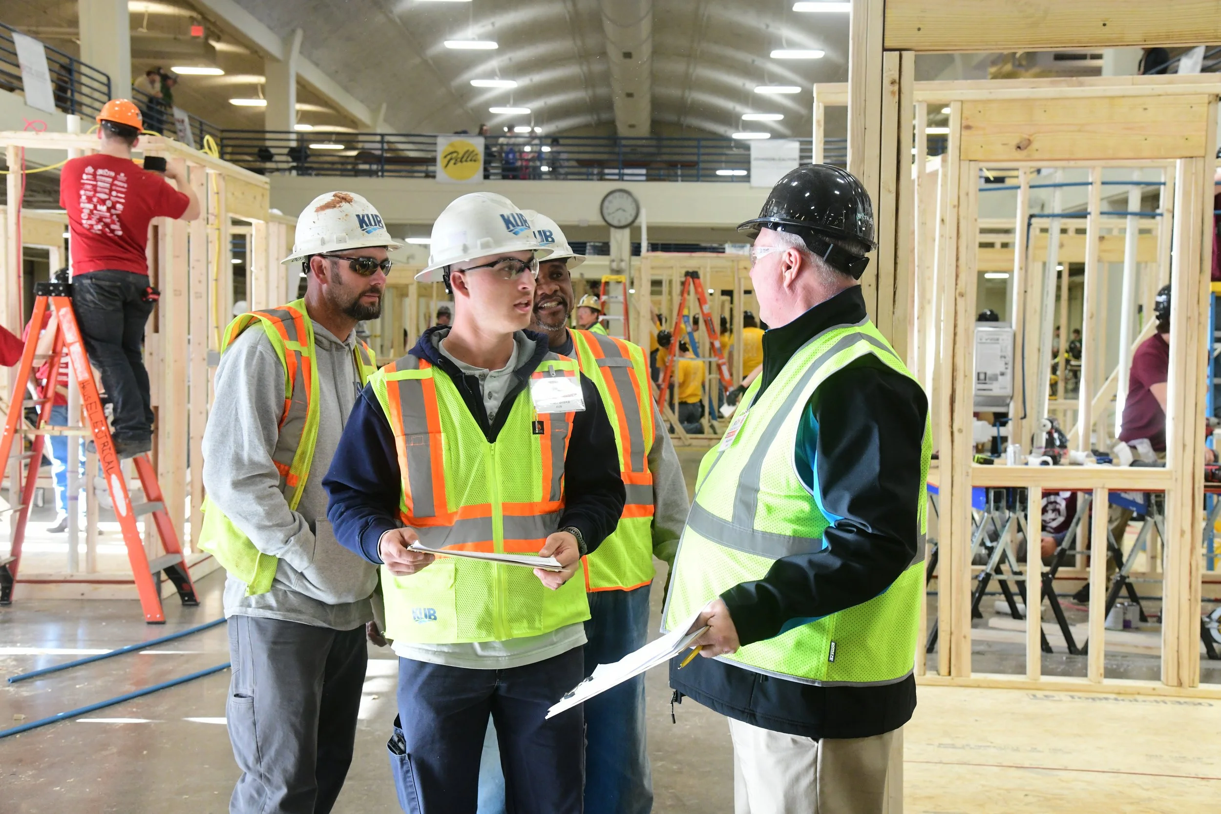 Construction workers and supervisors wearing helmets and safety vests inside a building under construction, discussing plans.
