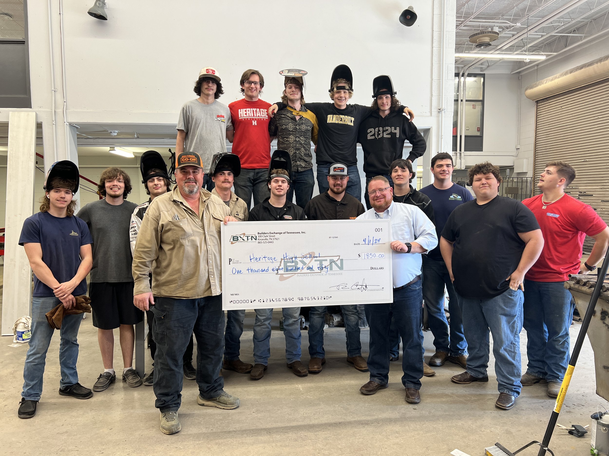 Group of high school students and adults posing together in a workshop, holding a large check for $1,850, with some students wearing welding helmets and safety gear.