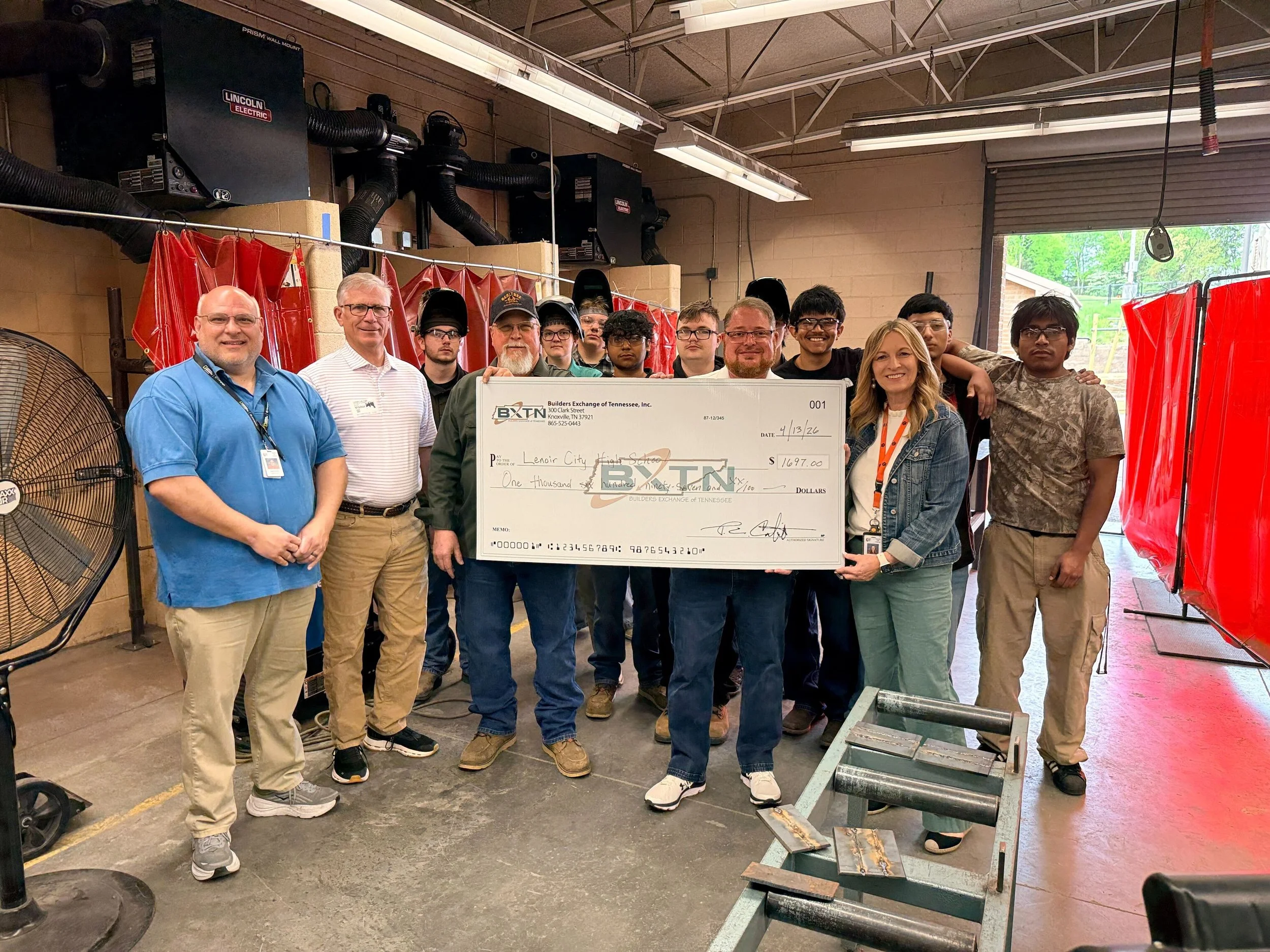 Group of people standing together inside a workshop or industrial space, holding a large check for $1,697.00, with tools and equipment around.