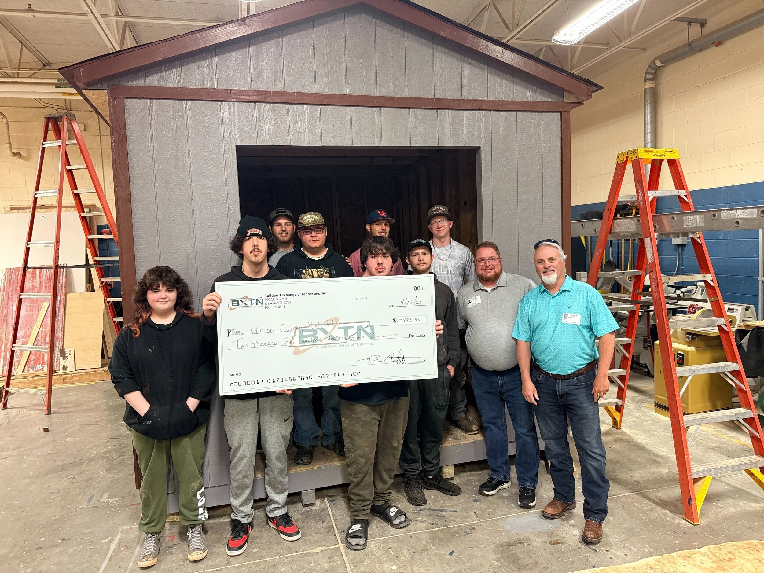 Group of people standing together inside a workshop with ladders and a small building structure. One person is holding a large check.