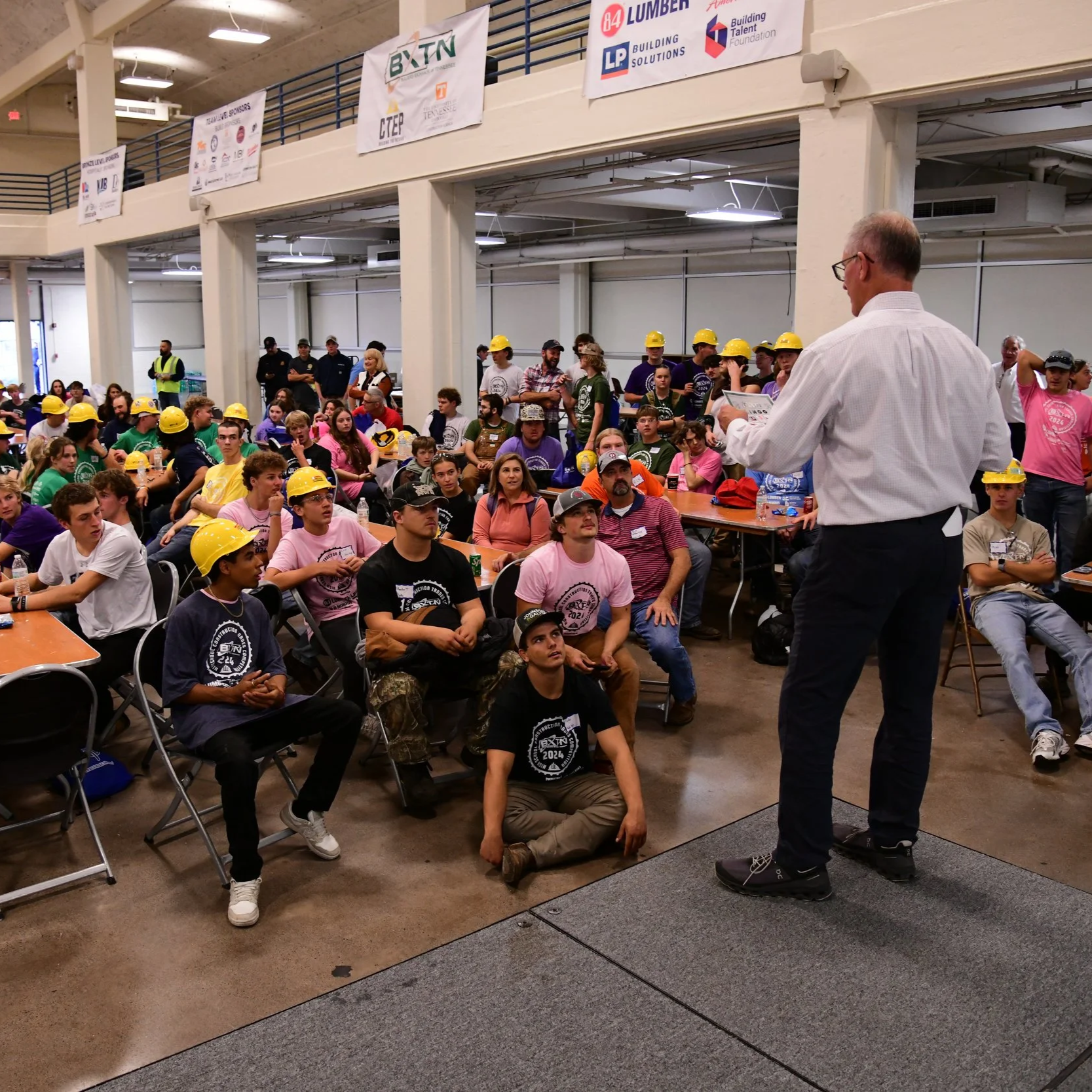 A group of young people wearing hard hats sitting and standing in an indoor conference or seminar room. An older man in a white shirt appears to be giving a presentation. There are banners hanging from the balcony above with various logos. Some of the attendees are paying attention, while others are looking around or talking.