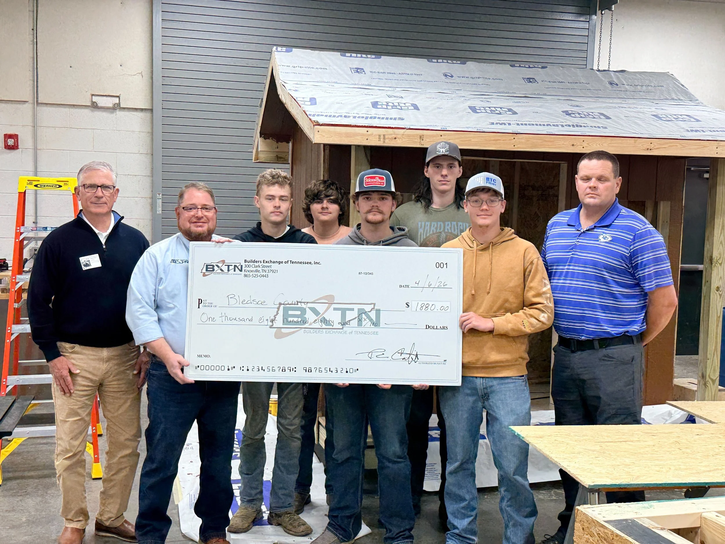 A group of six people standing inside a building, holding a large check. The check is made out to Bledsoe County for one thousand eight hundred eighty dollars. There are construction materials and a partially built wooden structure behind them.