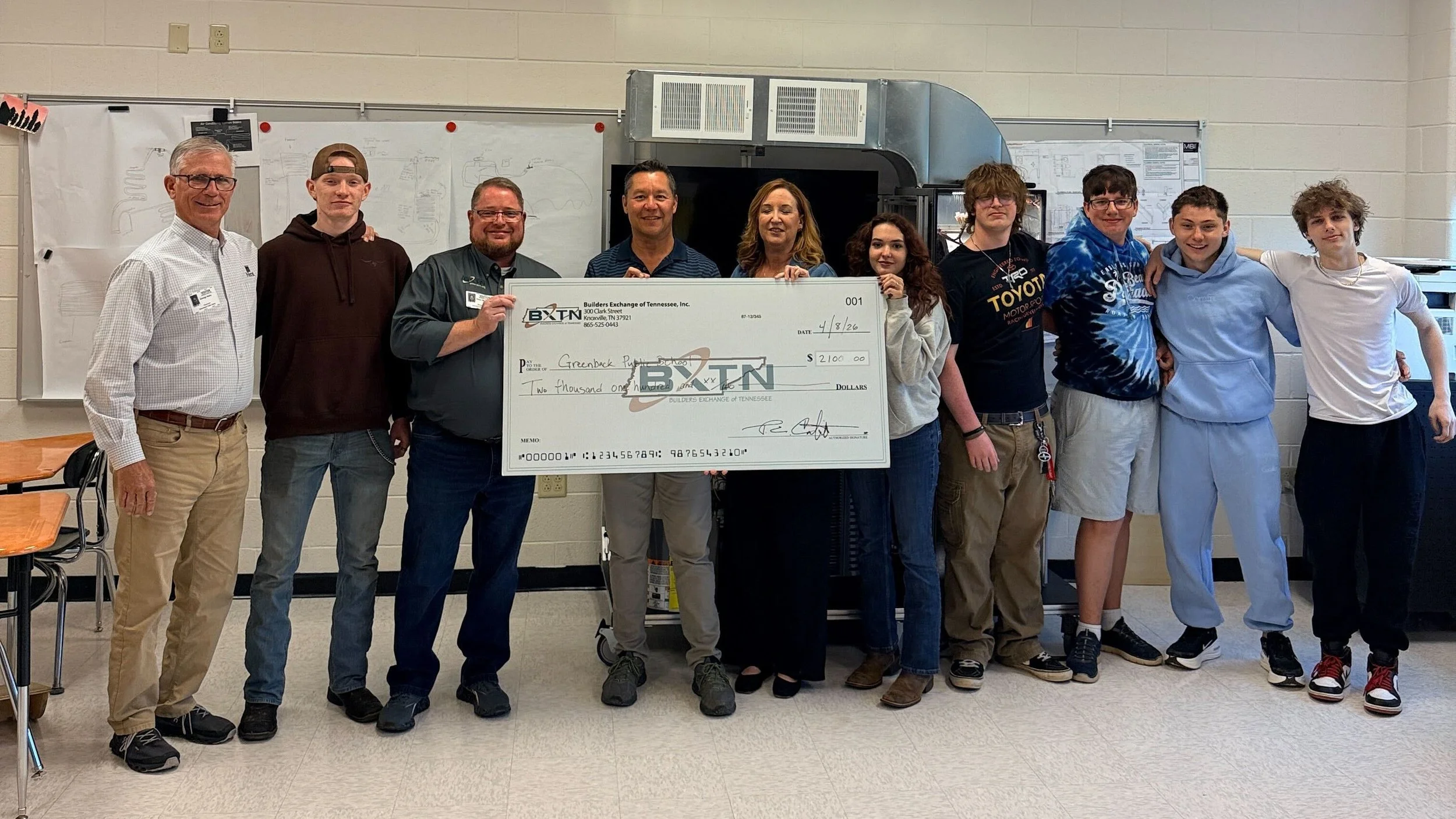 A group of ten people standing together in a classroom, holding an oversized check made out to Greenback Packaging. The group includes adults and teenagers, with some smiling at the camera. The classroom has white walls, desks, and a whiteboard with some drawings or notes.