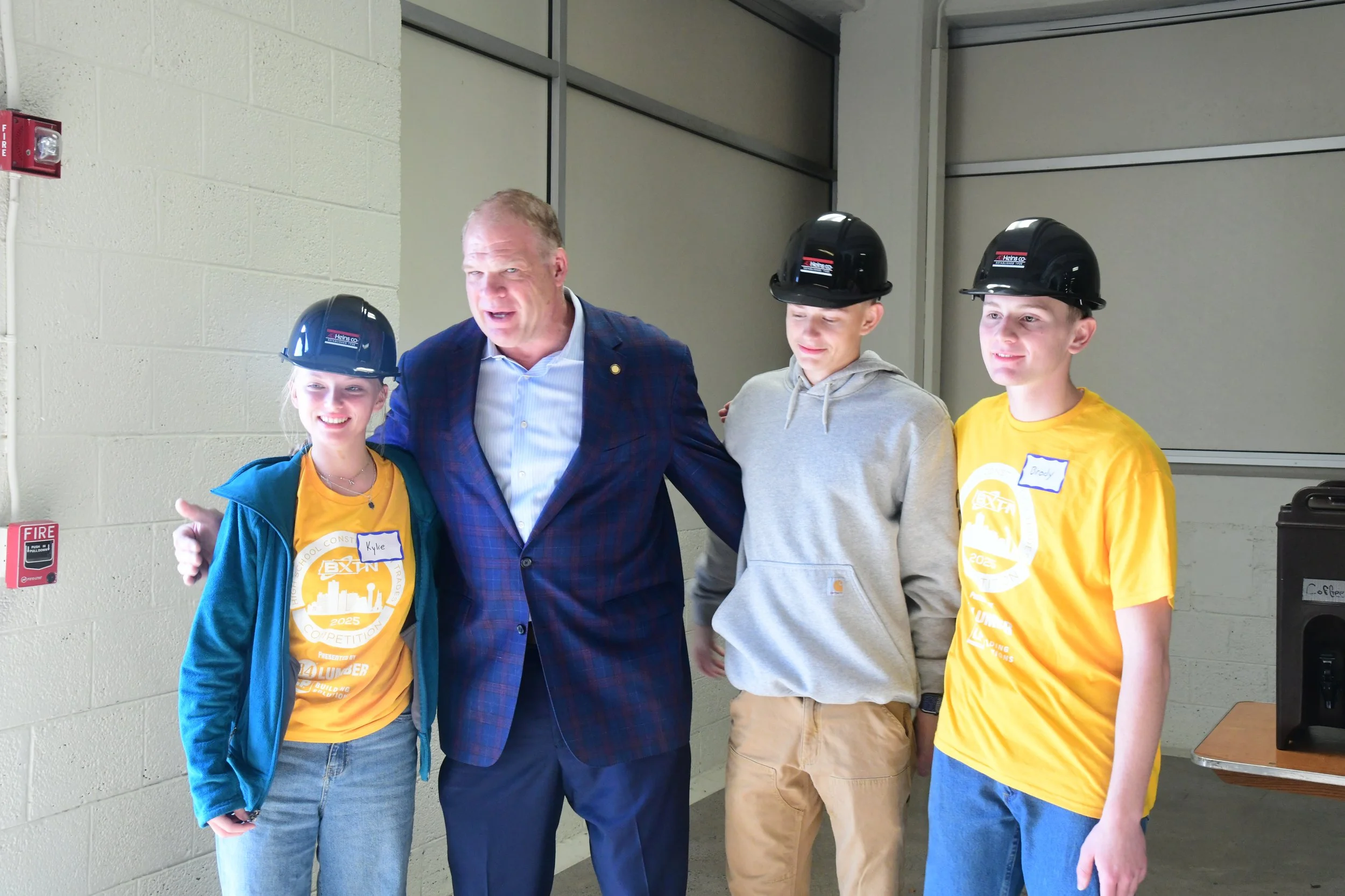 A man in a blue plaid suit with three teenagers, all wearing safety helmets, inside an industrial or construction building. The teenagers are wearing yellow and gray clothing and appear to be part of a construction or safety event.