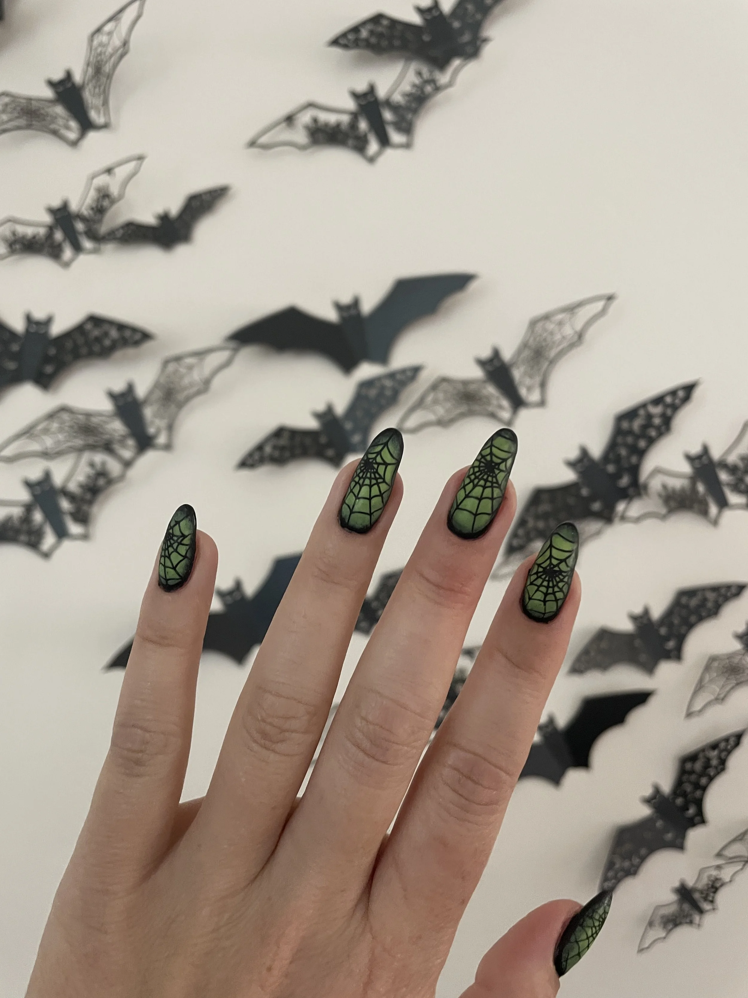 A hand with green Halloween-themed nails featuring web and spider designs, in front of a white surface decorated with black paper bats, some with lace-like patterns.