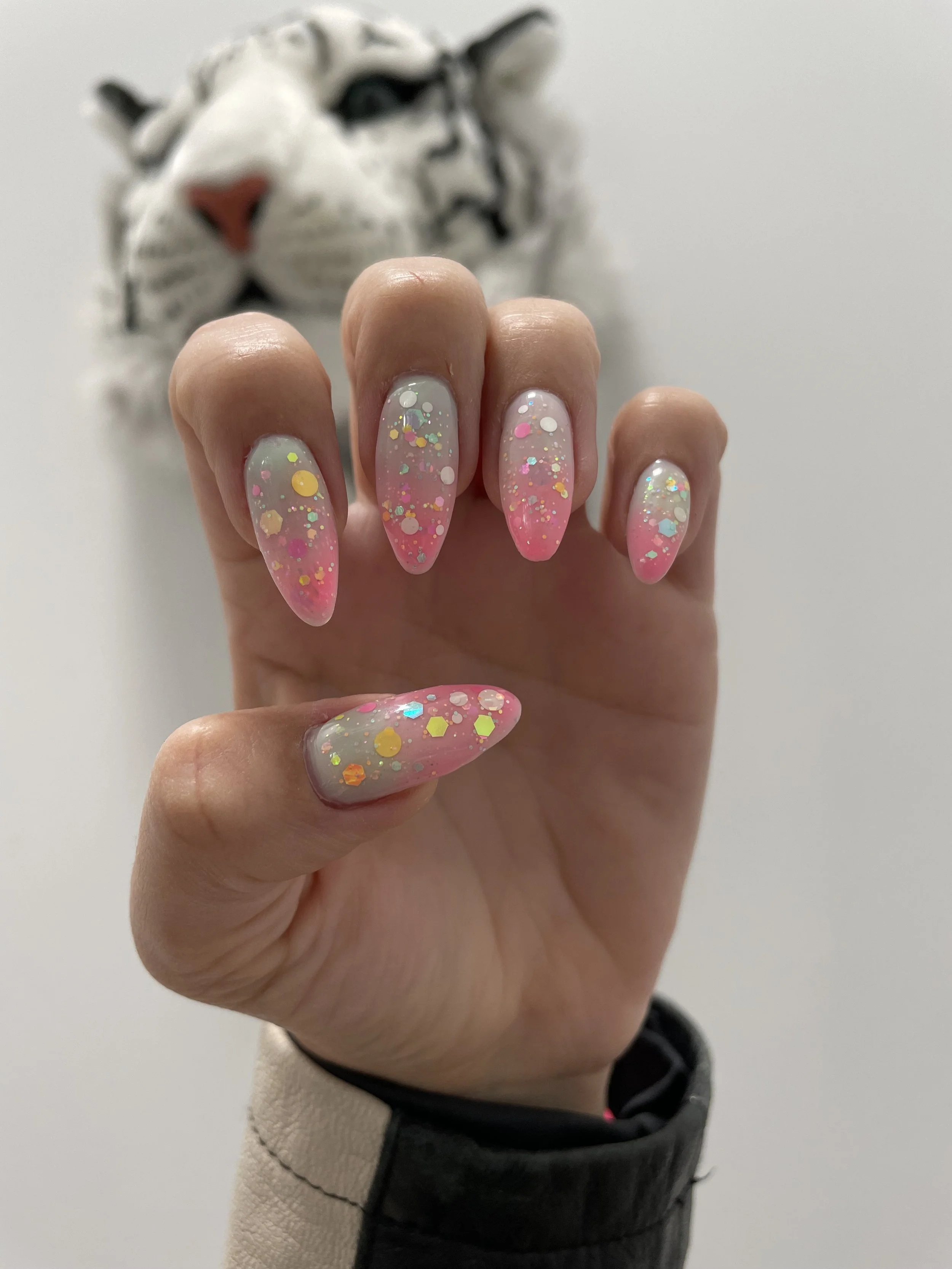Close-up of a hand with long, almond-shaped nails painted with clear gel and decorated with multicolored glitter, in front of a blurry white tiger statue.