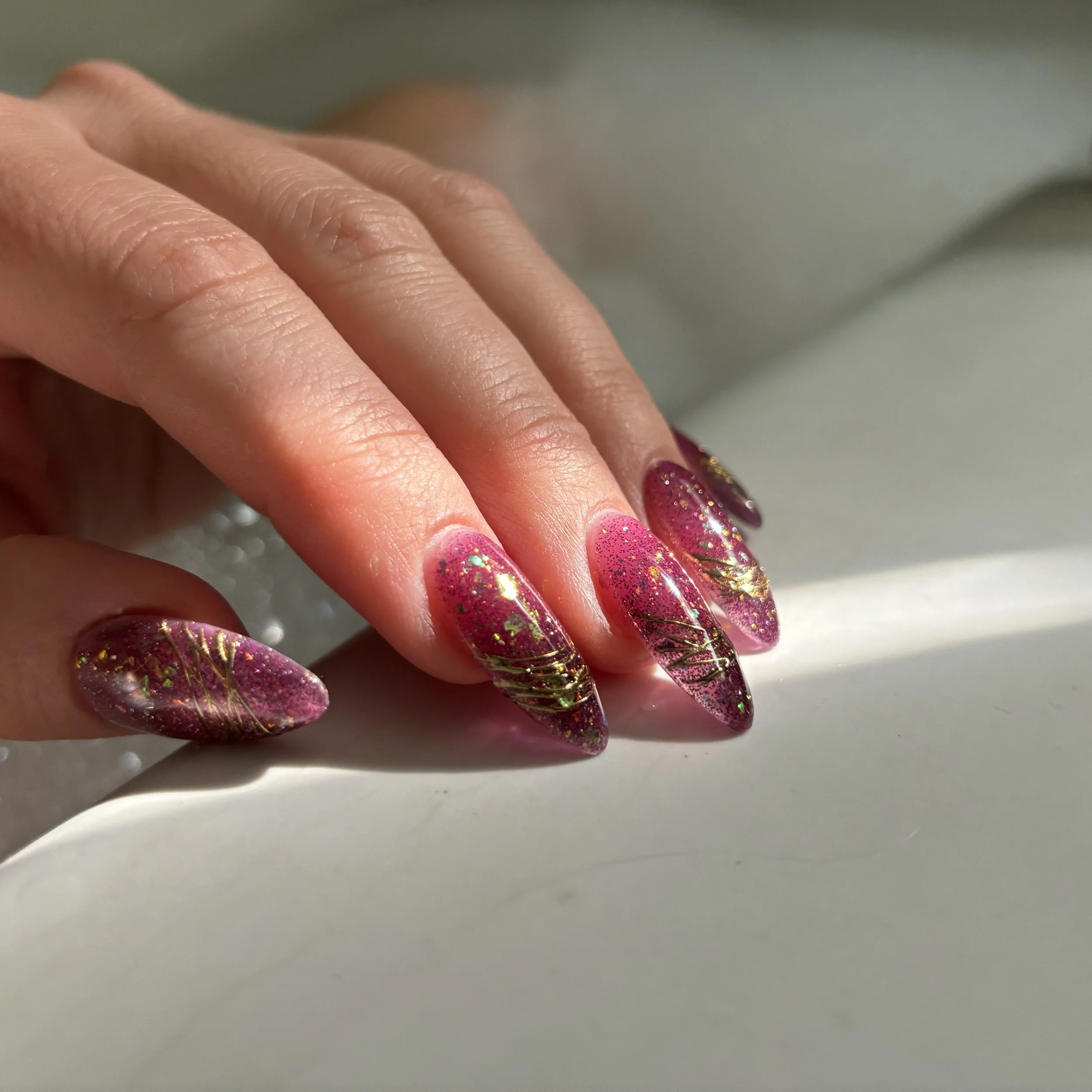 Close-up of a person's hand with decorated nails, featuring pink polish with glitter and gold accents, on a white surface.