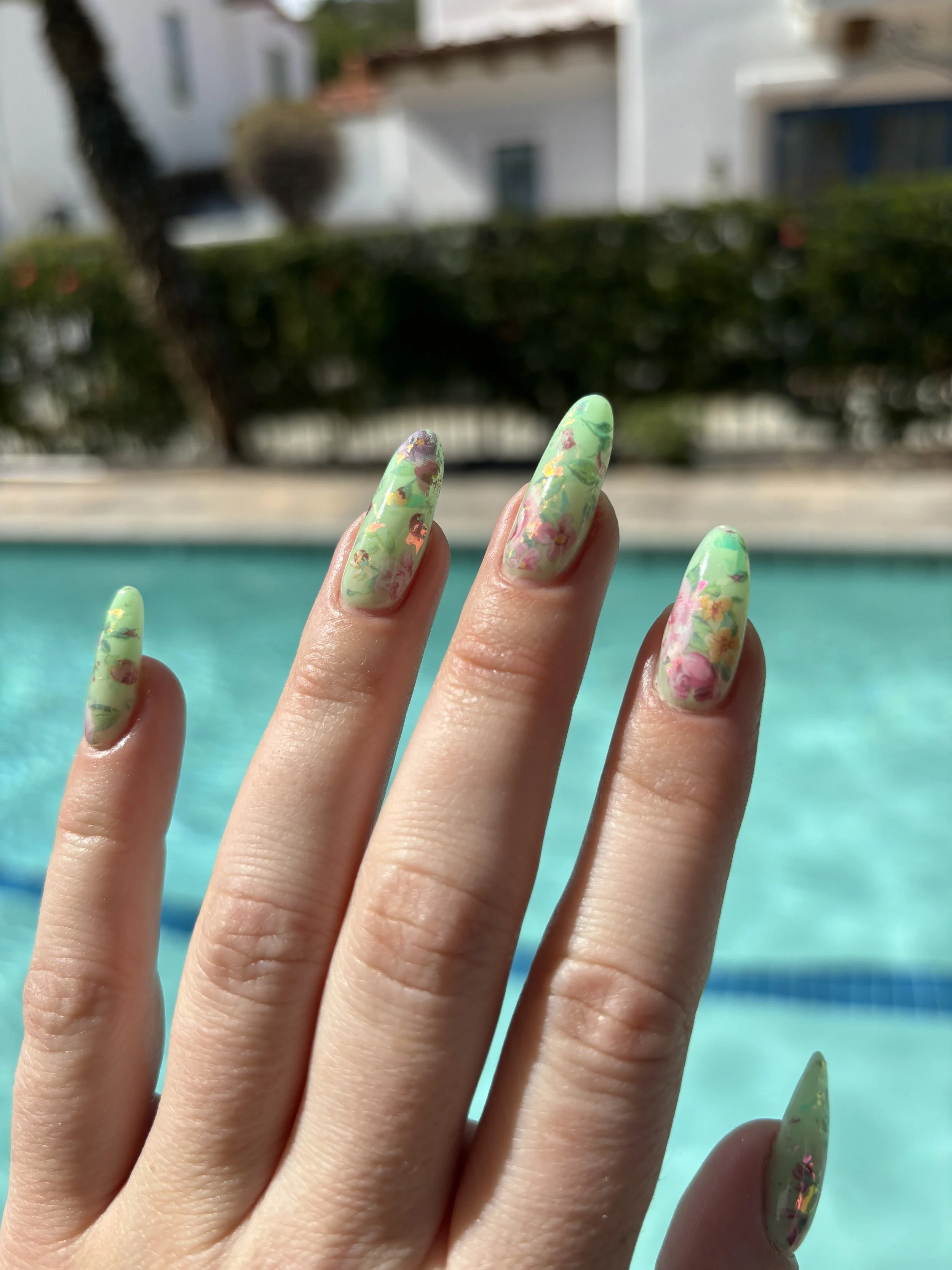 Close-up of a hand with long, decorated nails in front of a swimming pool, with houses and trees in the background.
