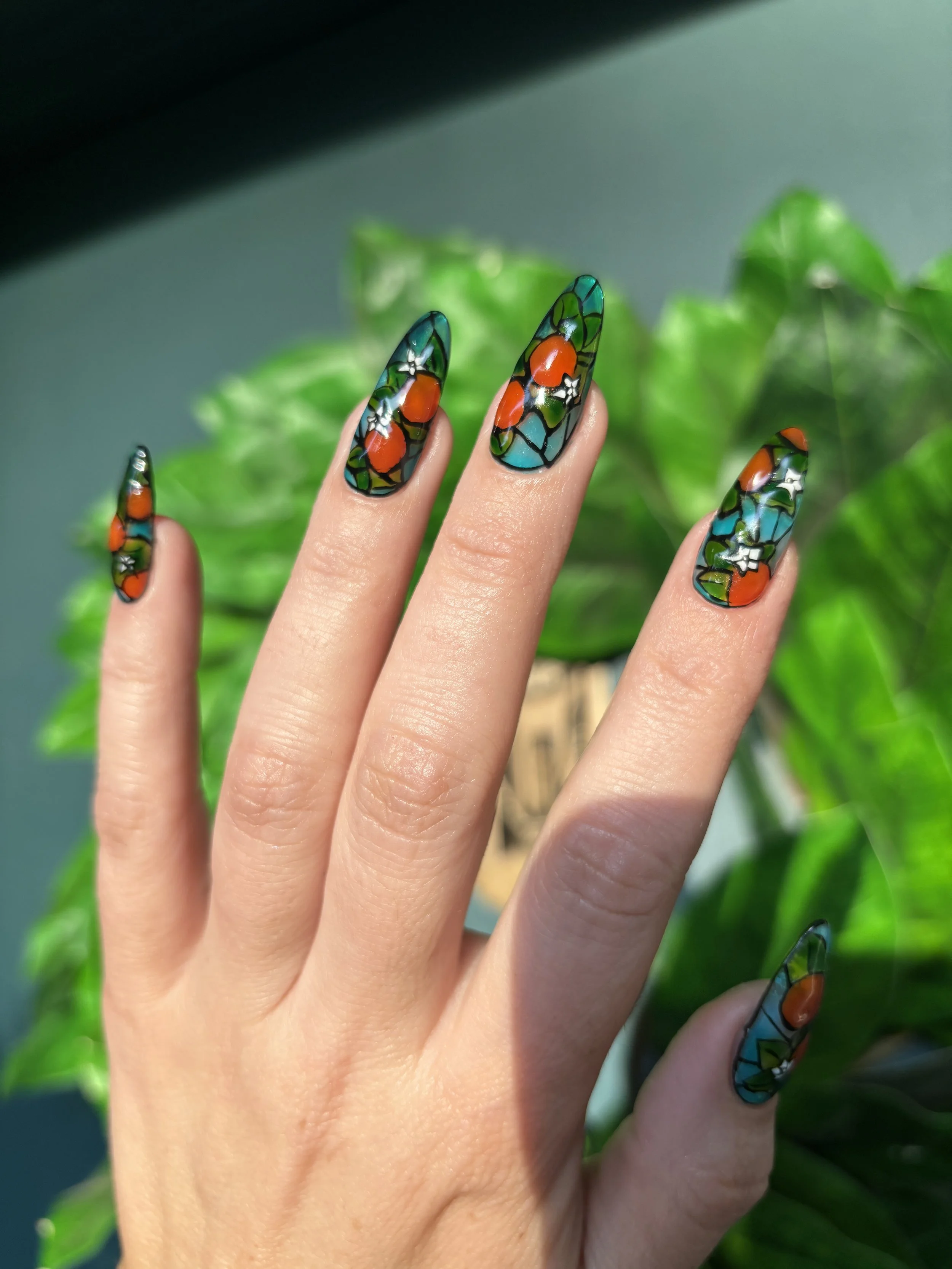 Hand with long, colorful, stained glass-style painted nails in front of green foliage.
