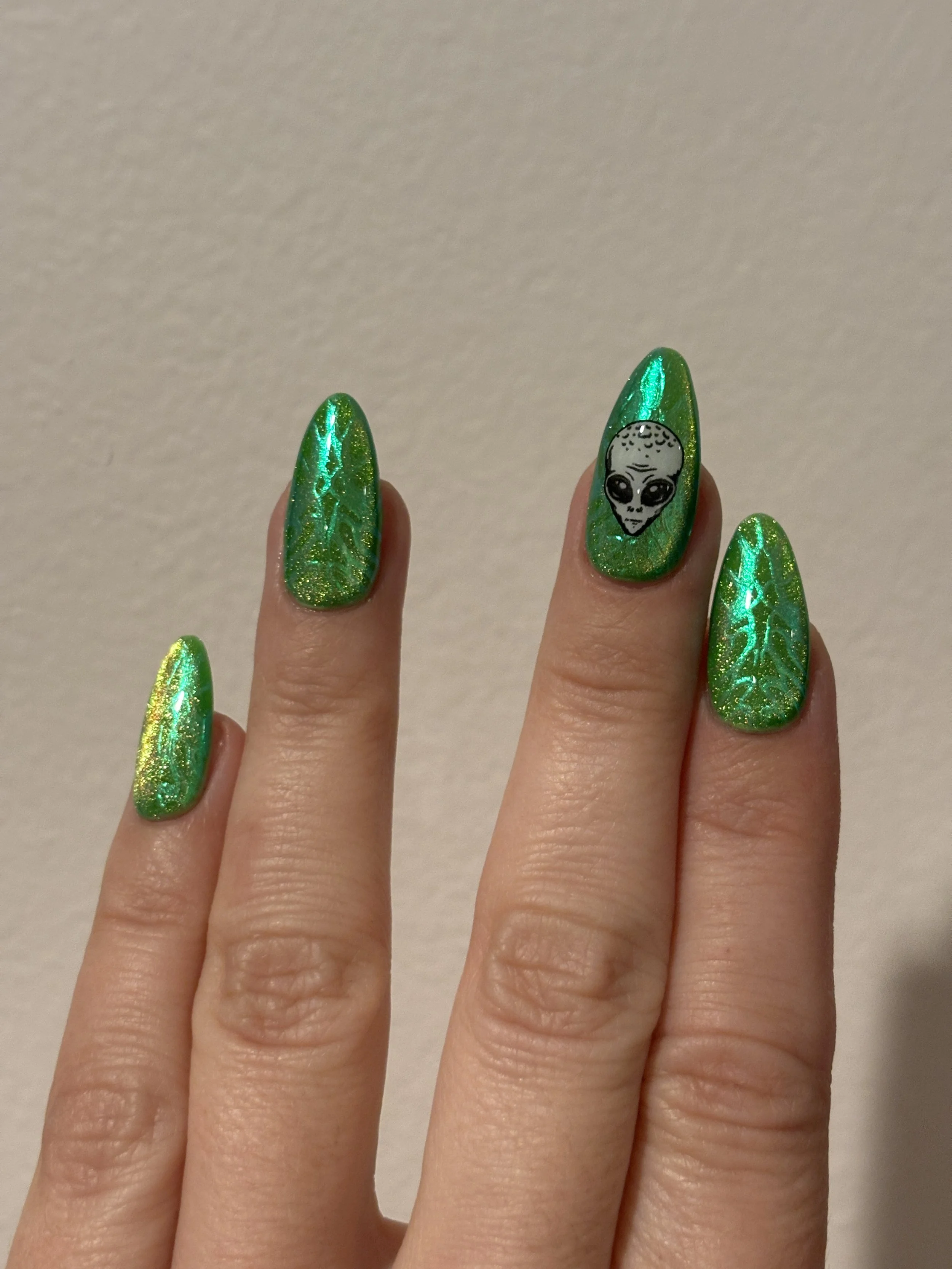 Hand with green holographic nail polish and an alien face sticker on one fingernail.