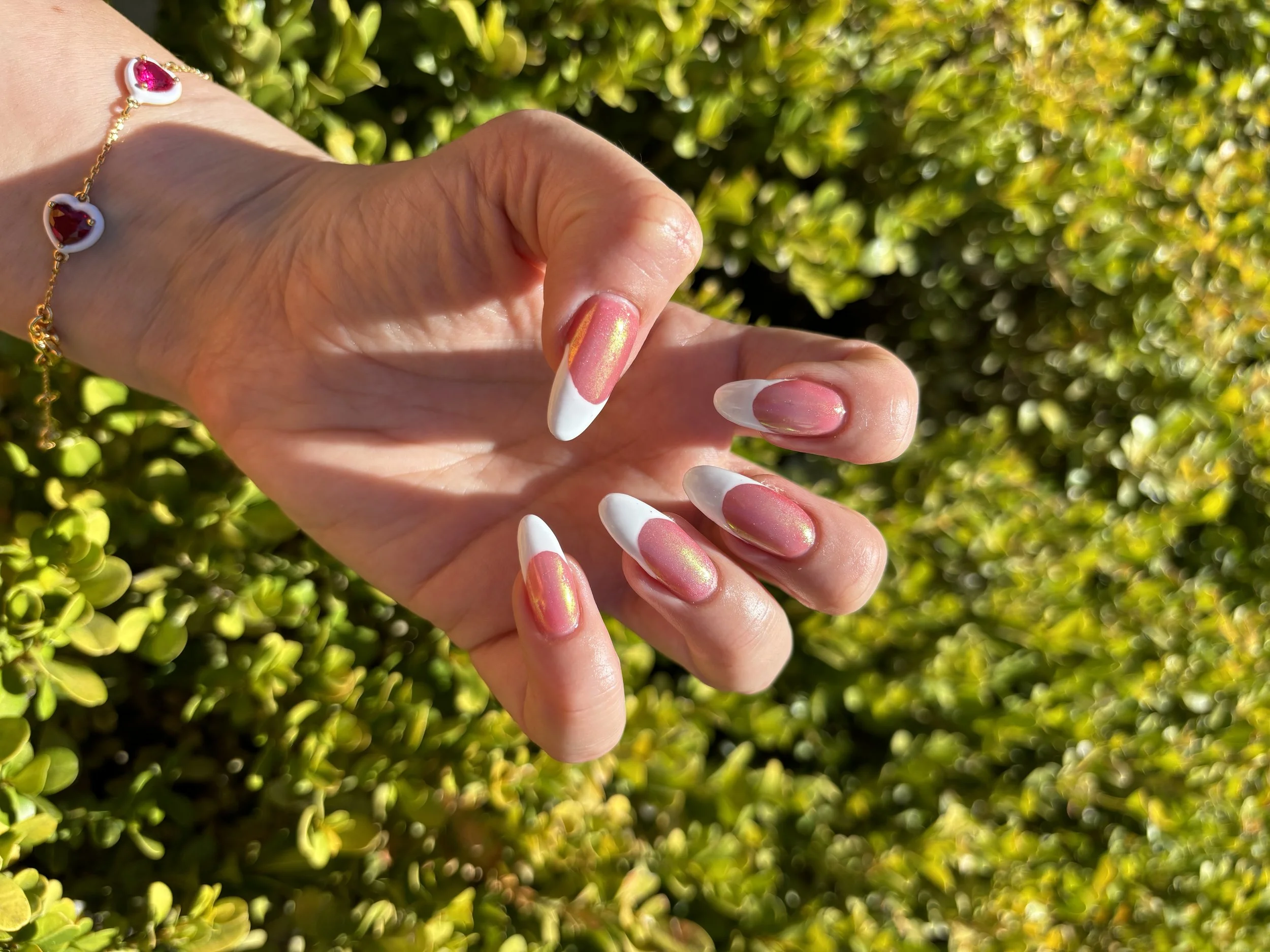 Close-up of a woman's hand with a gold bracelet featuring red and pink heart-shaped stones, extended fingers with a French manicure, against a background of green foliage.