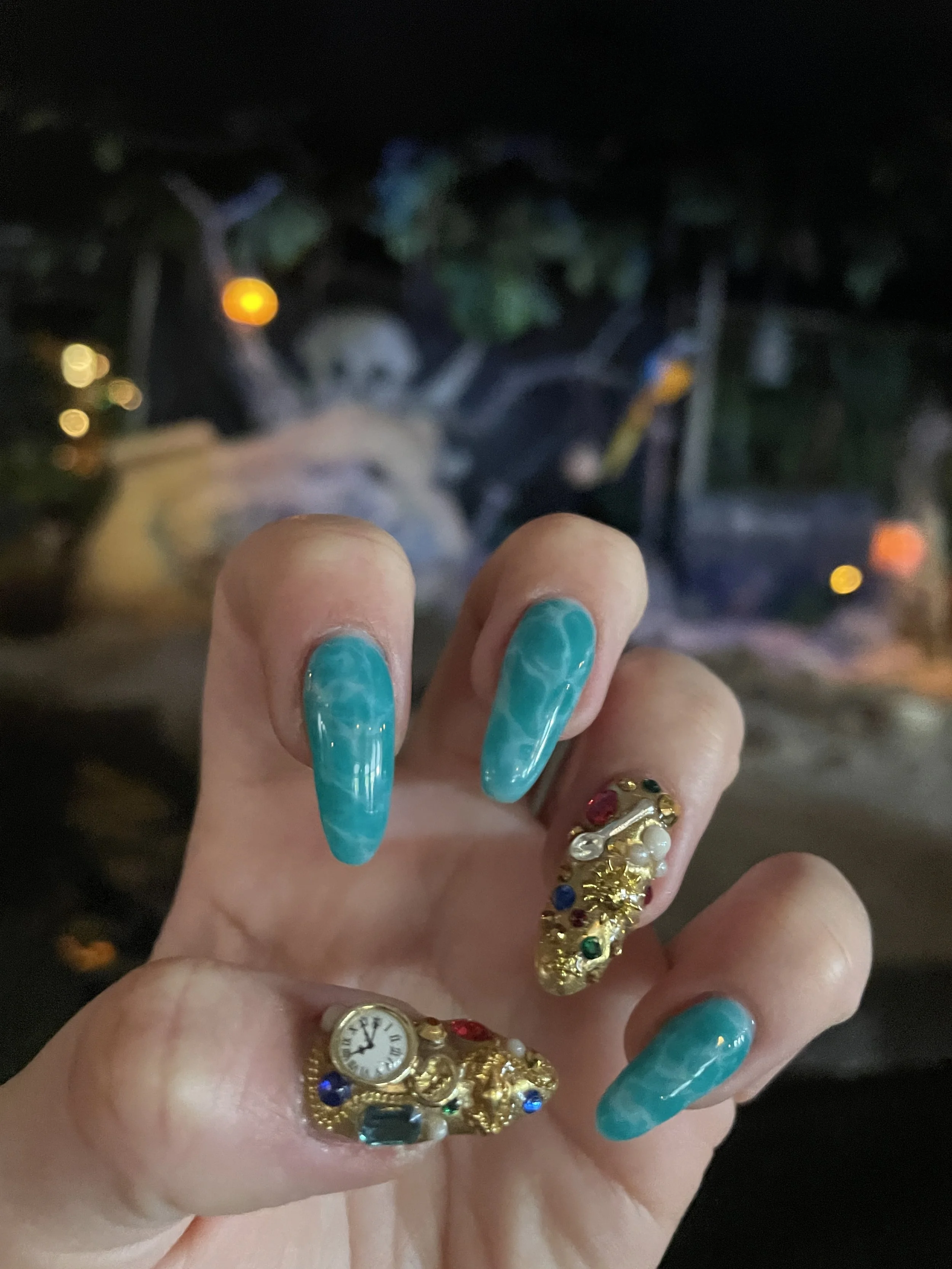 Close-up of a person's hand with turquoise-colored long nails and rings, including a gold ring with a clock face and a ring with multicolored rhinestones, against a blurry night street background with streetlights.