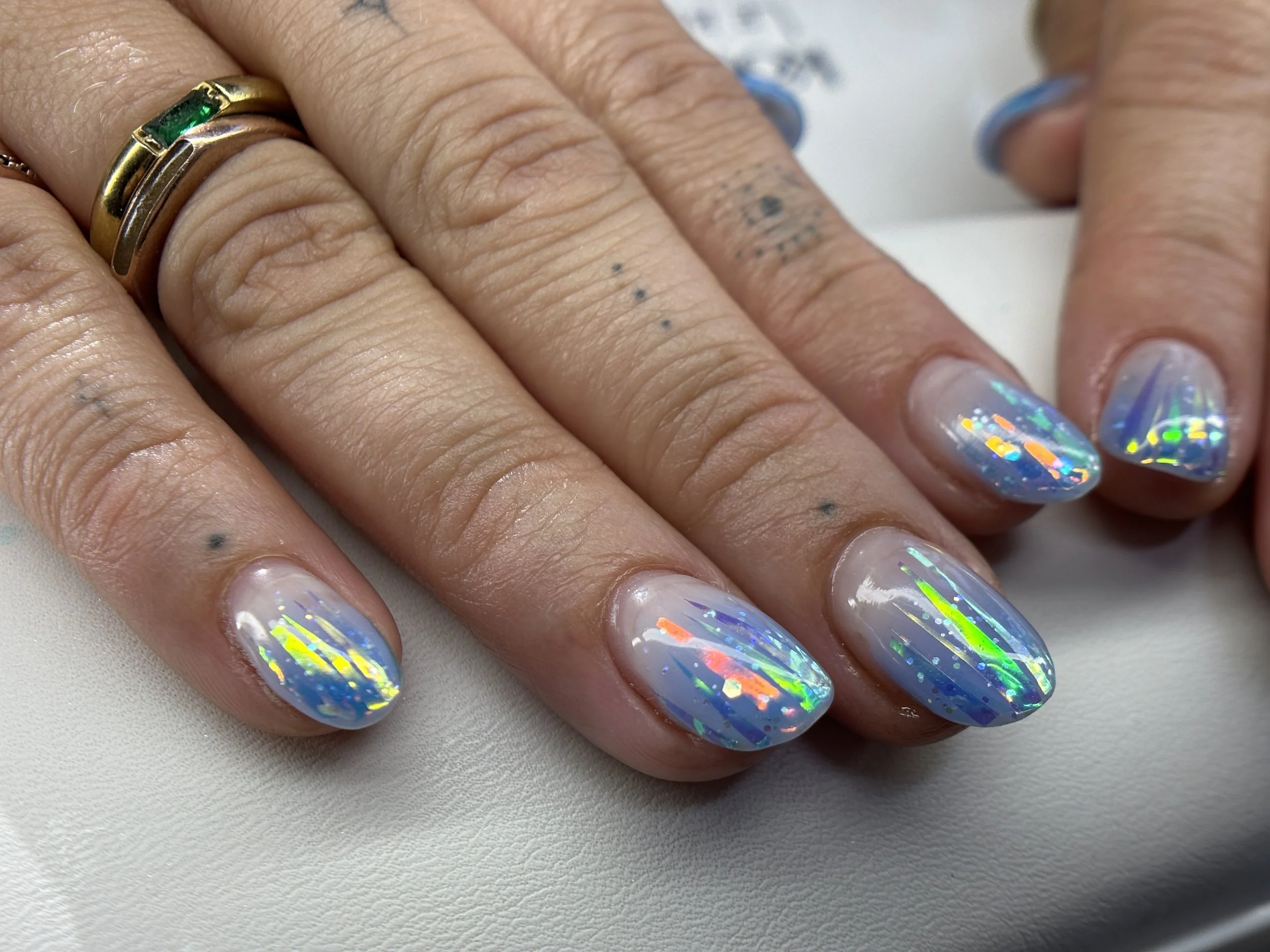 Close-up image of a person's hand showing holographic, iridescent nail polish on their nails with a red, green, and silver reflection. The person is wearing multiple rings including one with a rectangular green gemstone, and has small black tattoos o