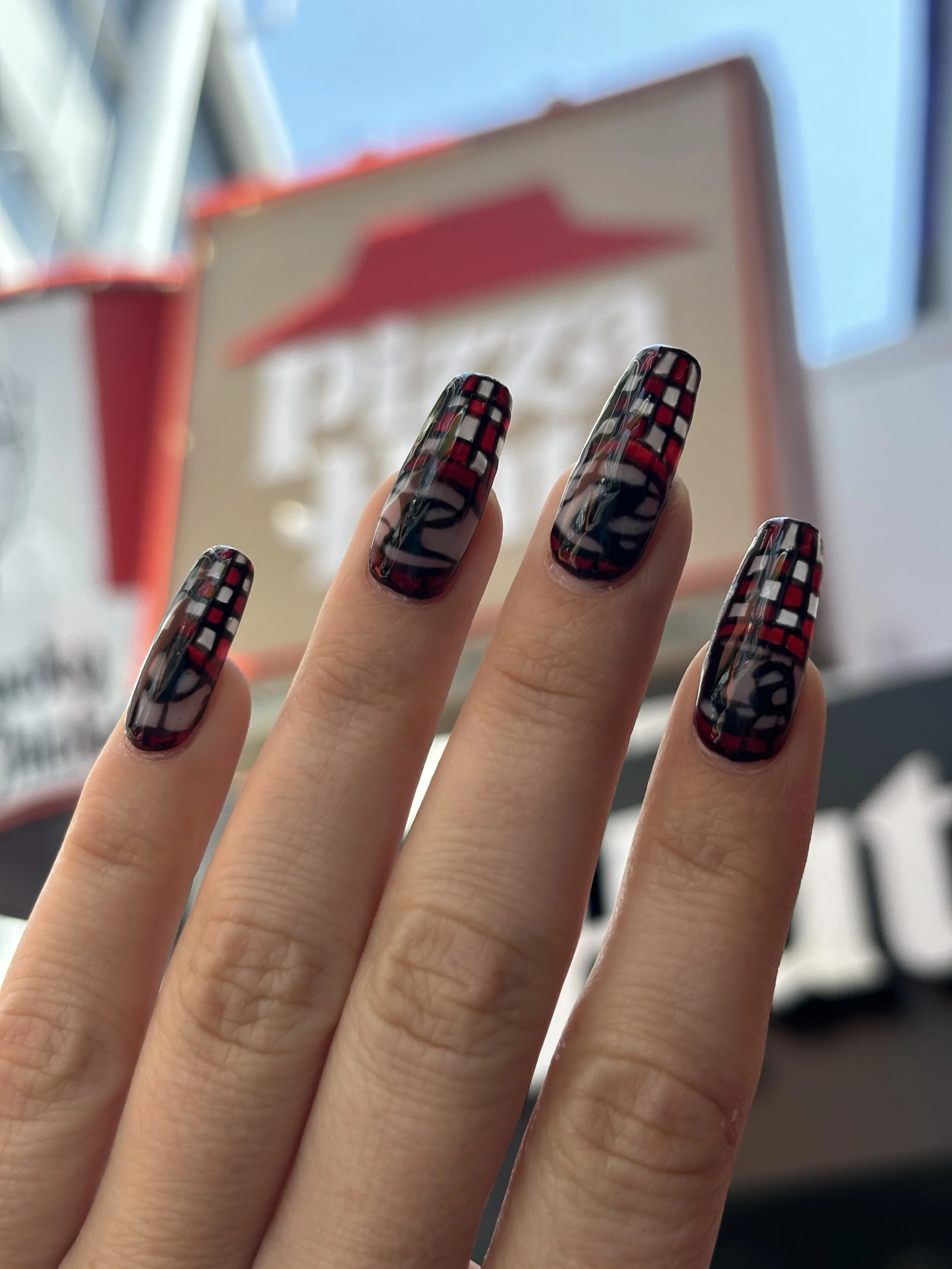 Close-up of a hand with long, checkered nails in black, red, and white, with blurring of background objects including a sign and metal structures.
