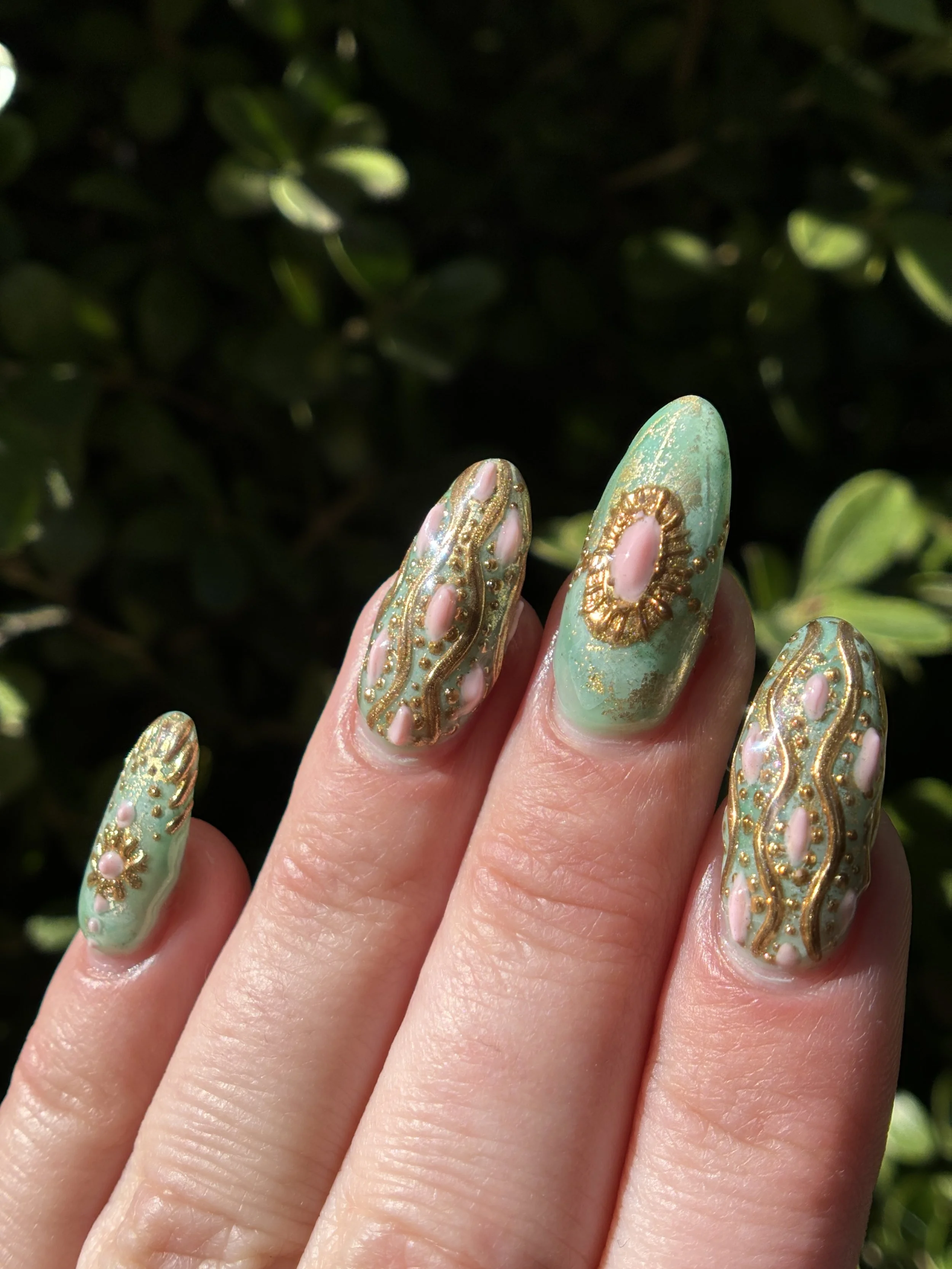 Close-up of a hand with decorated nails featuring a mint green base with gold accents and small white pearls, held against a backdrop of green leaves.