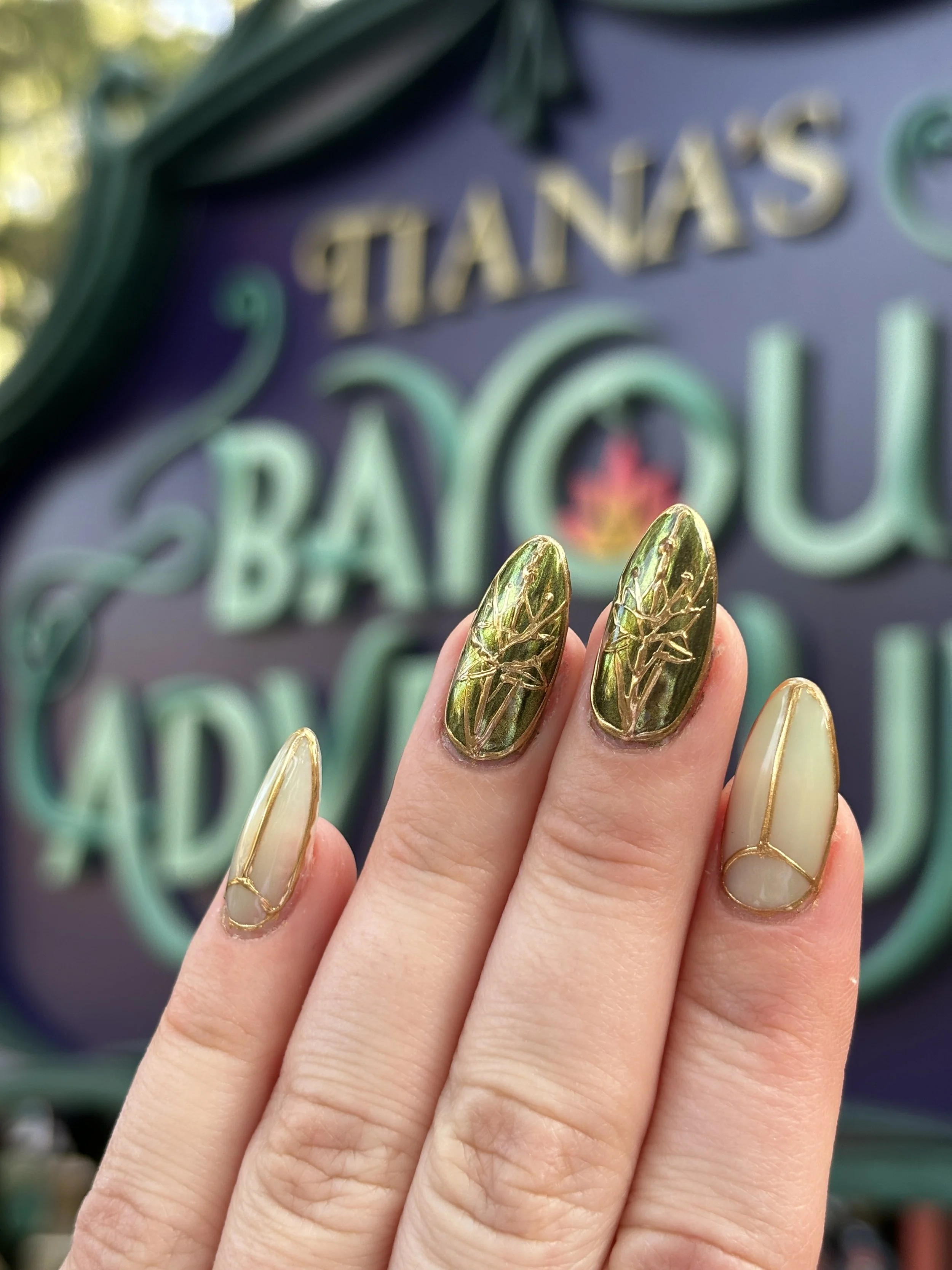 Close-up of a person's hand with decorative gold and green nails, featuring leaf patterns, in front of a blurred background with a sign reading 'TANAS'.