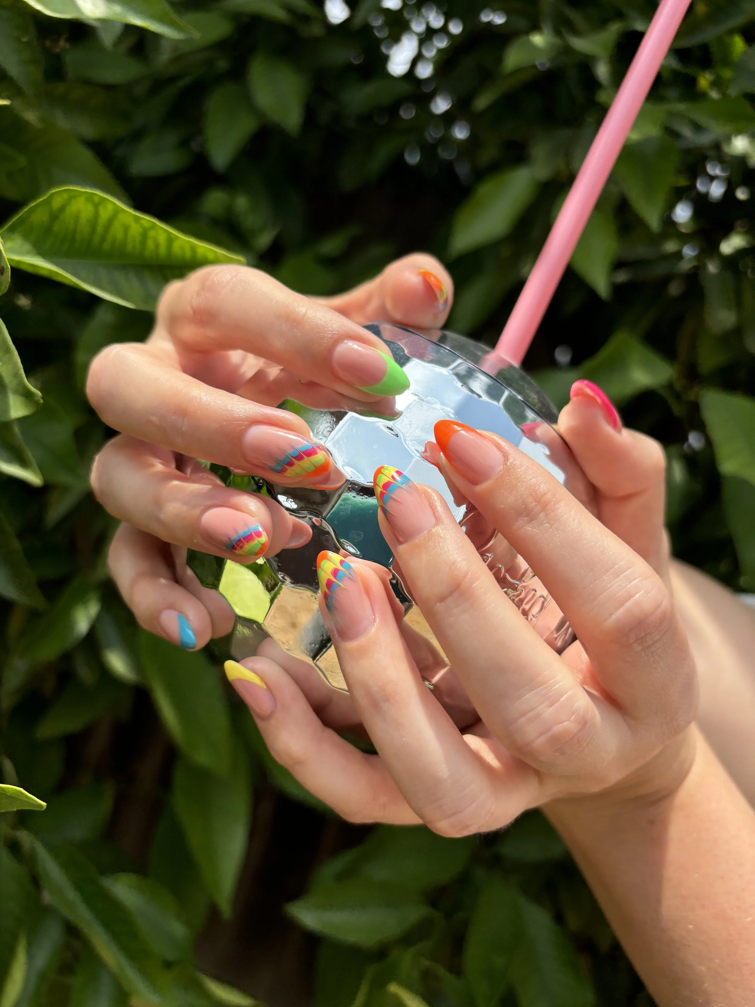 Close-up of hands holding a clear shiny crystal with colorful rainbow nails in front of green leafy background, with pink straw visible.