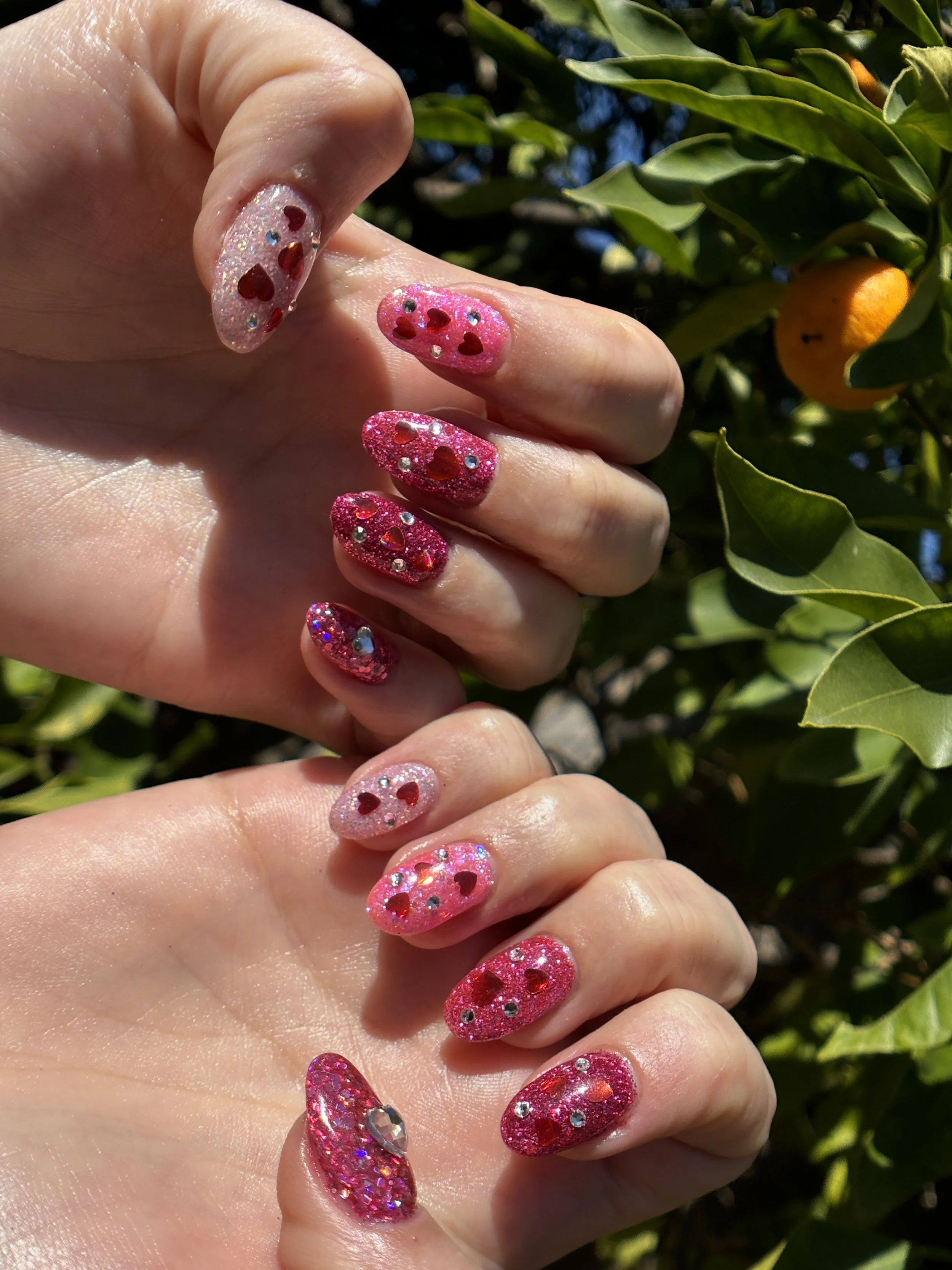 Close-up of a hand with decorated nails featuring pink and white glitter nail polish with red heart-shaped and small round rhinestone accents, set against green leaves with small orange fruit.
