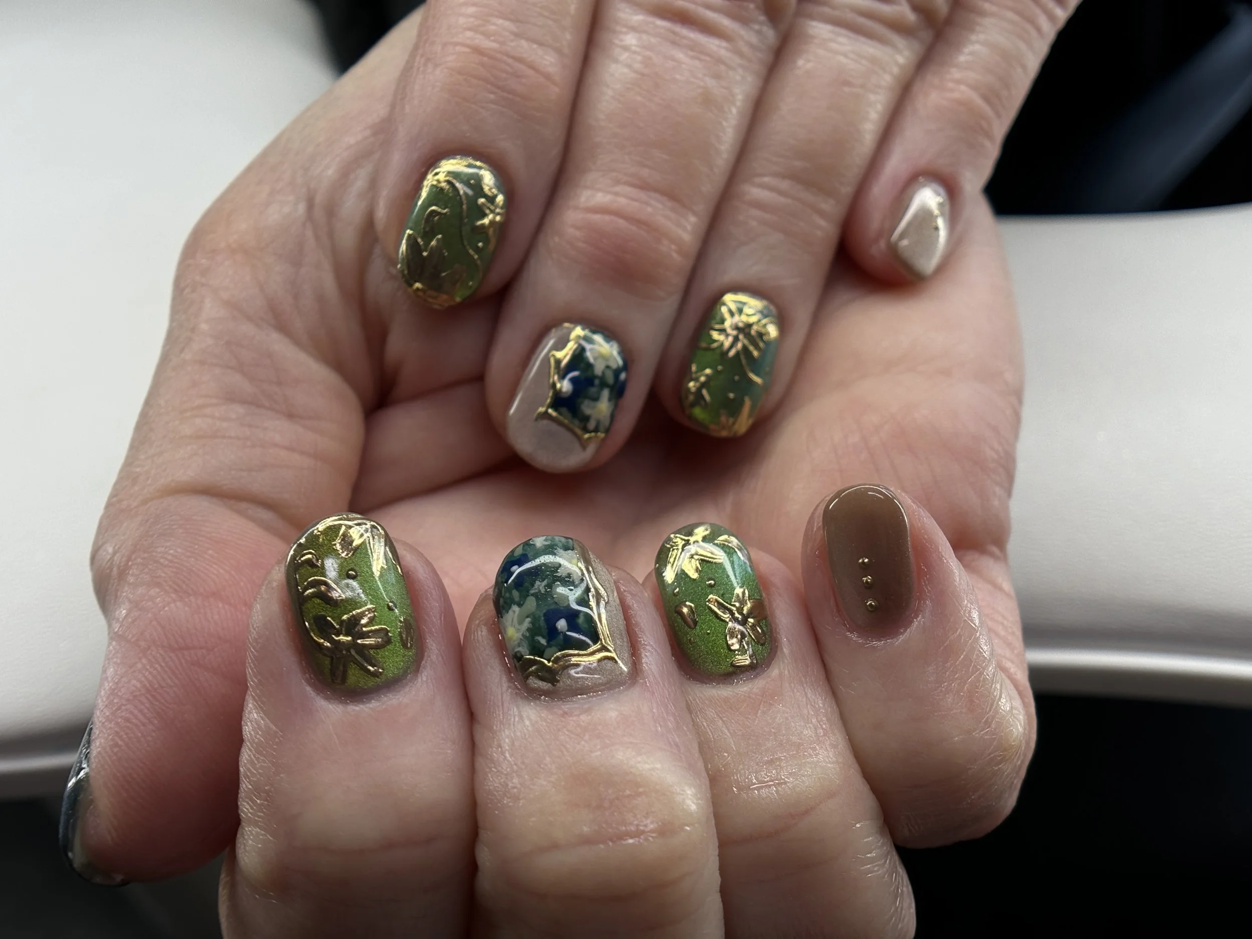 A hand with manicured nails painted in shades of green and beige, featuring gold leaf designs and decorative patterns.