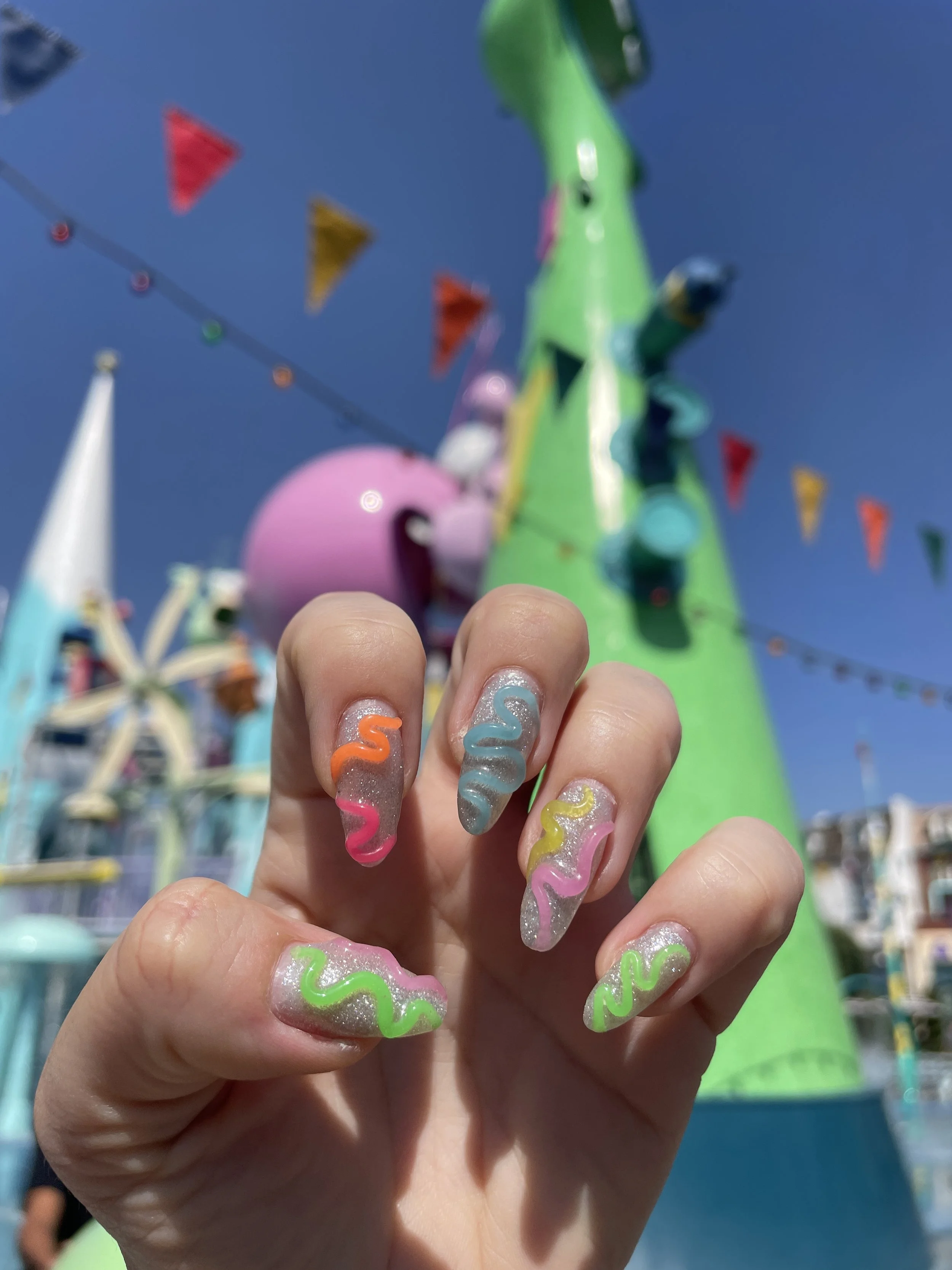 Close-up of a hand with colorful, curly, glittery nail art, held in front of a carnival or amusement park ride with bright colors and bunting flags.