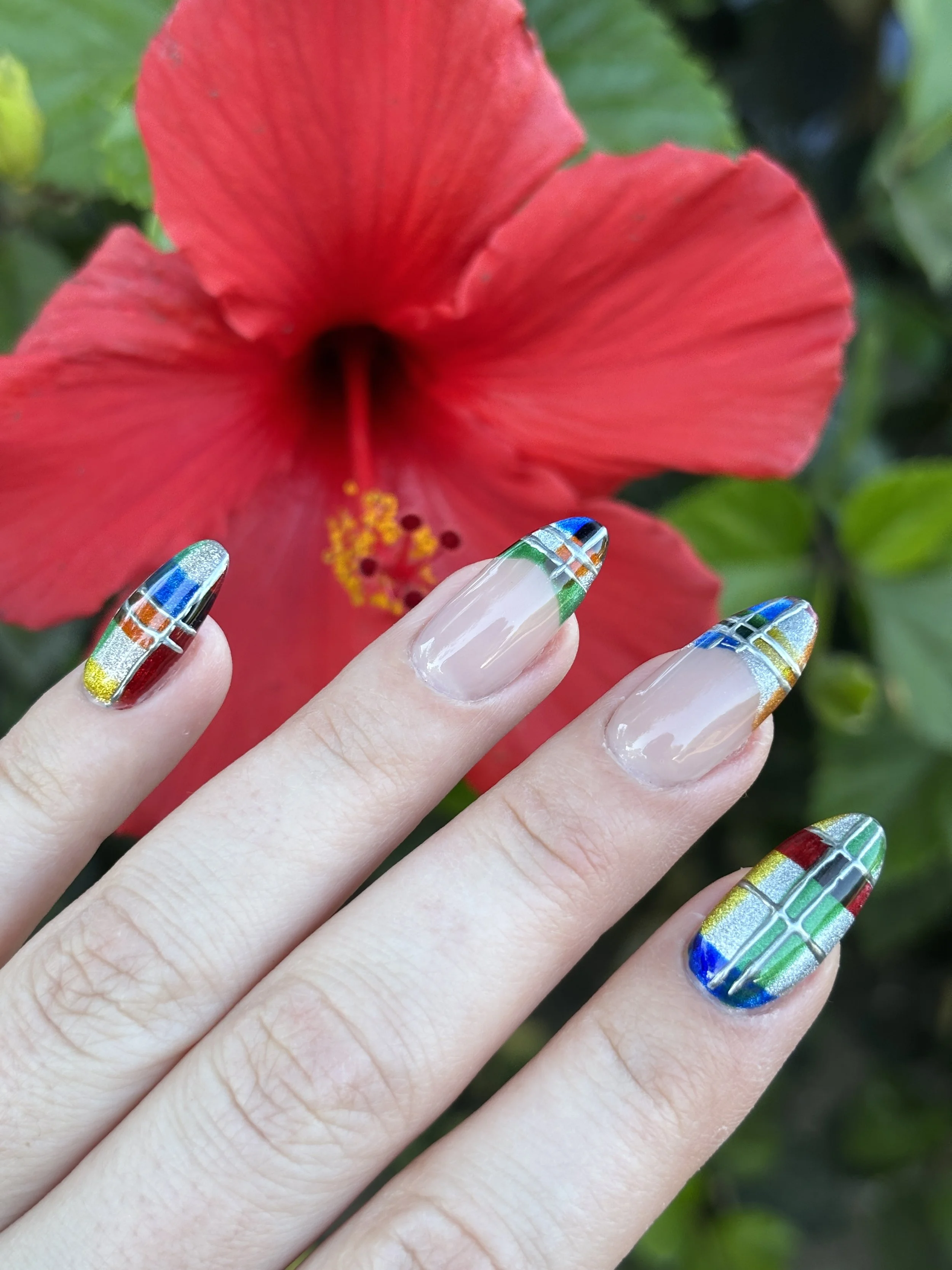 Hand with colorful, geometric patterned nail art in front of a red hibiscus flower with green leaves background.