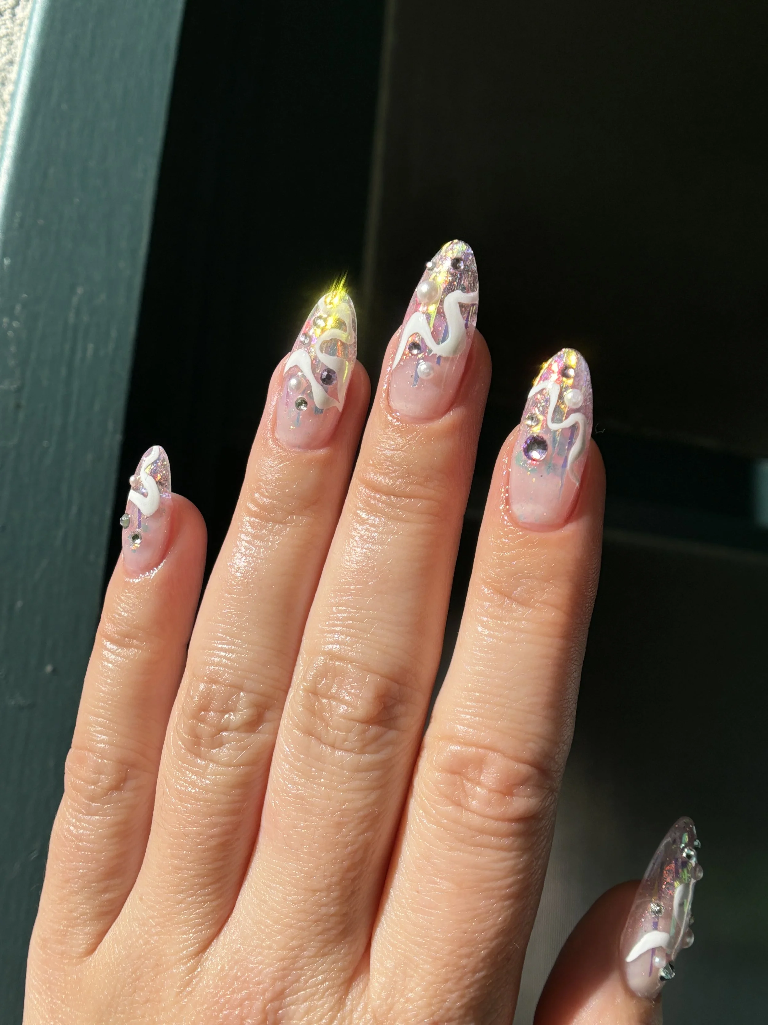 Close-up of a hand displaying decorated fingernails with holographic, iridescent nail polish and white swirl designs, adorned with small rhinestones.