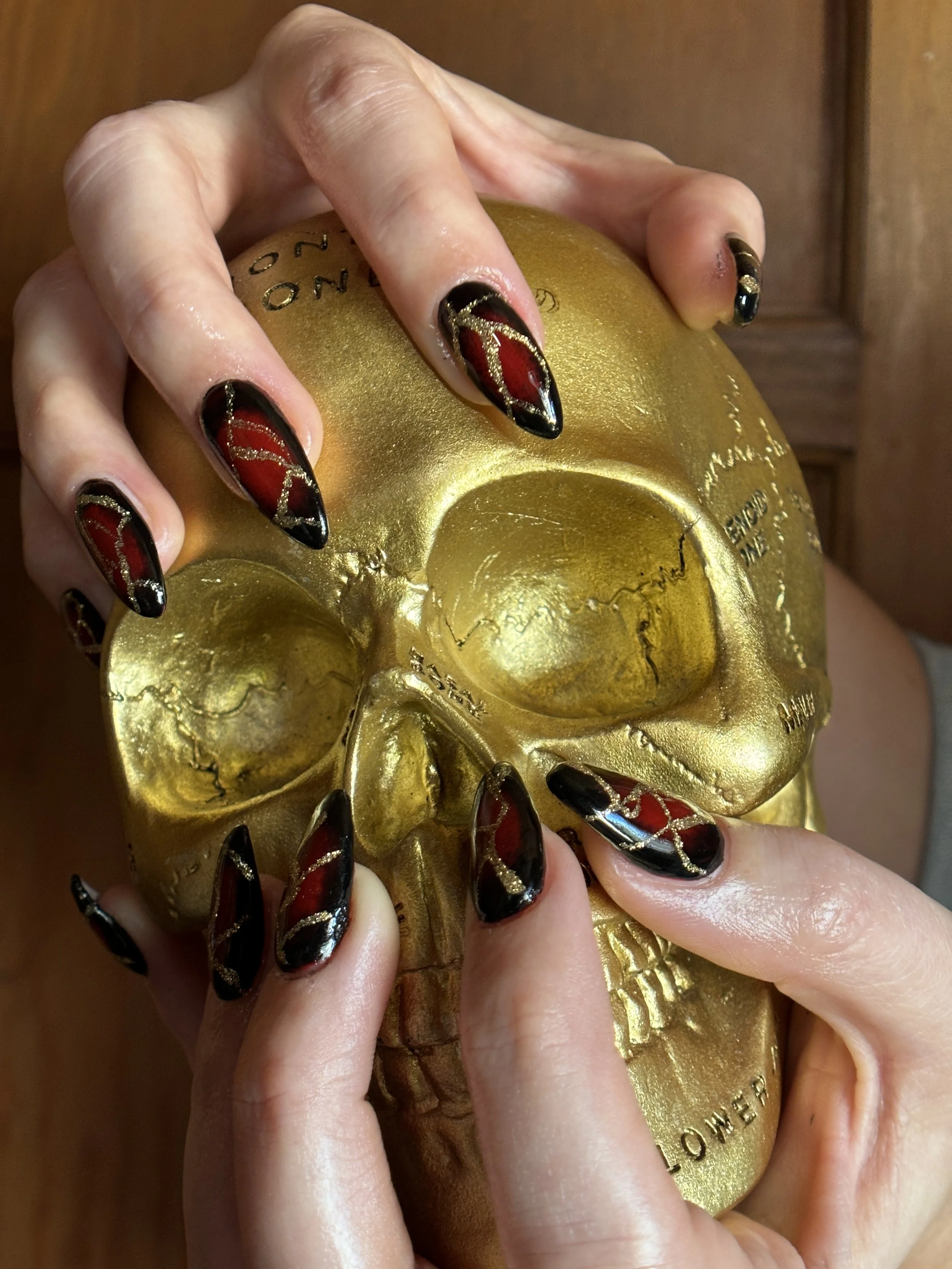 A hand with intricately designed black, red, and gold nail polish holding a gold-colored skull ornament.