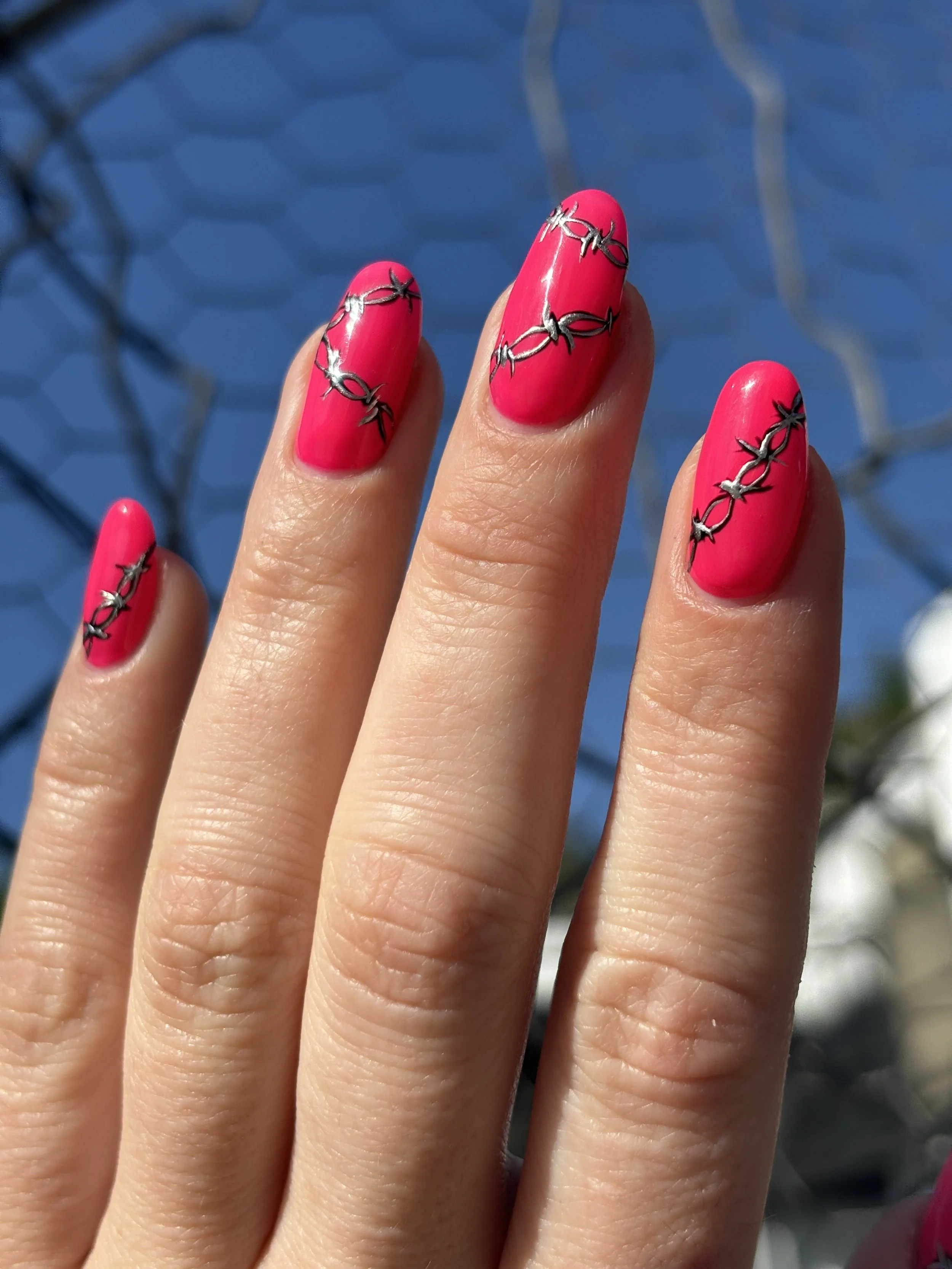 Close-up of three fingers with bright pink nail polish and silver thorns painted around the nails, against a blurred blue background.