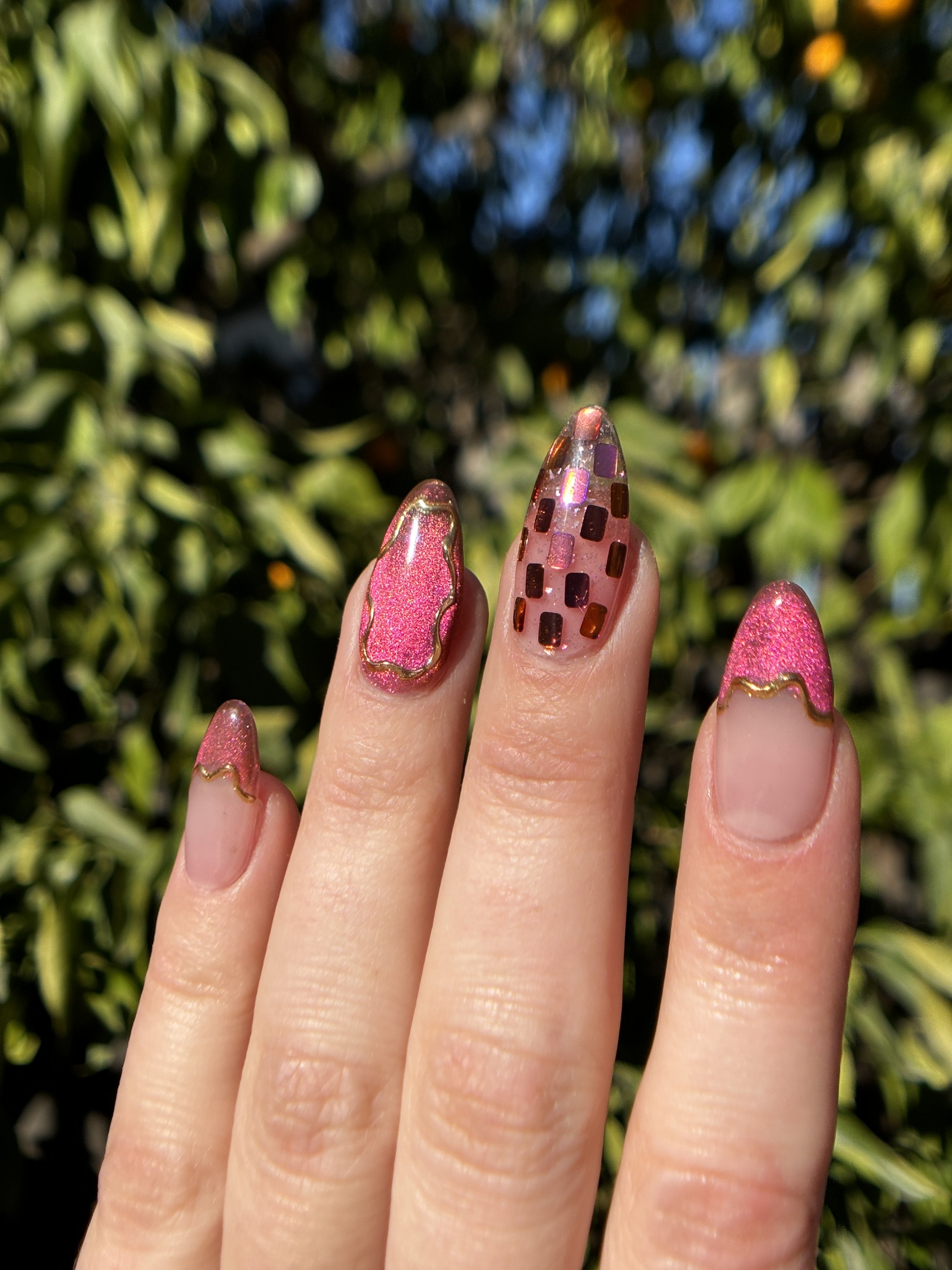 Close-up of four fingers with decorated nails, held against a background of green foliage. The nails feature pink, glittery, and patterned designs.