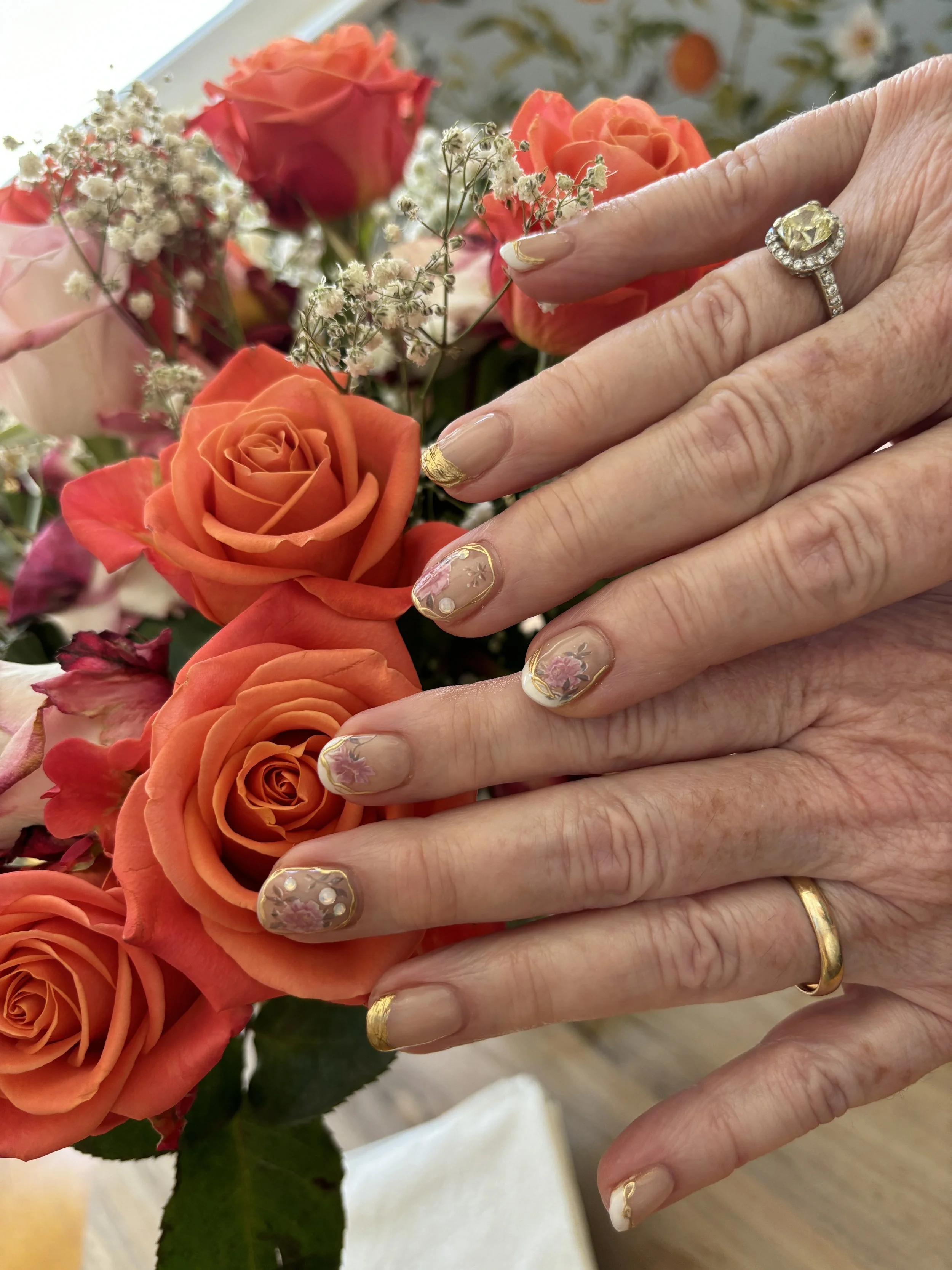 Close-up of a person's hand with decorated nails wearing two rings, one with a large yellow gemstone and the other a gold band, resting on an arrangement of orange and pink roses and white baby's breath flowers.
