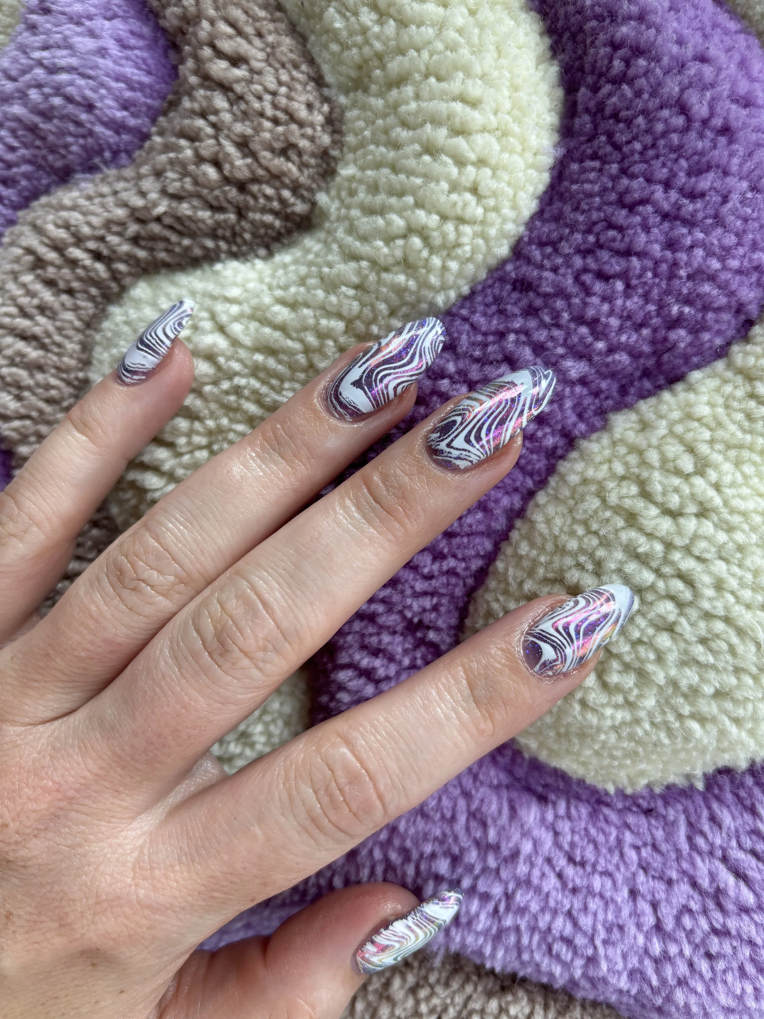 Close-up of a hand with nails featuring holographic, rainbow-colored, marbled nail art against a background of a multicolored, textured knit blanket in purple, cream, brown, and beige.