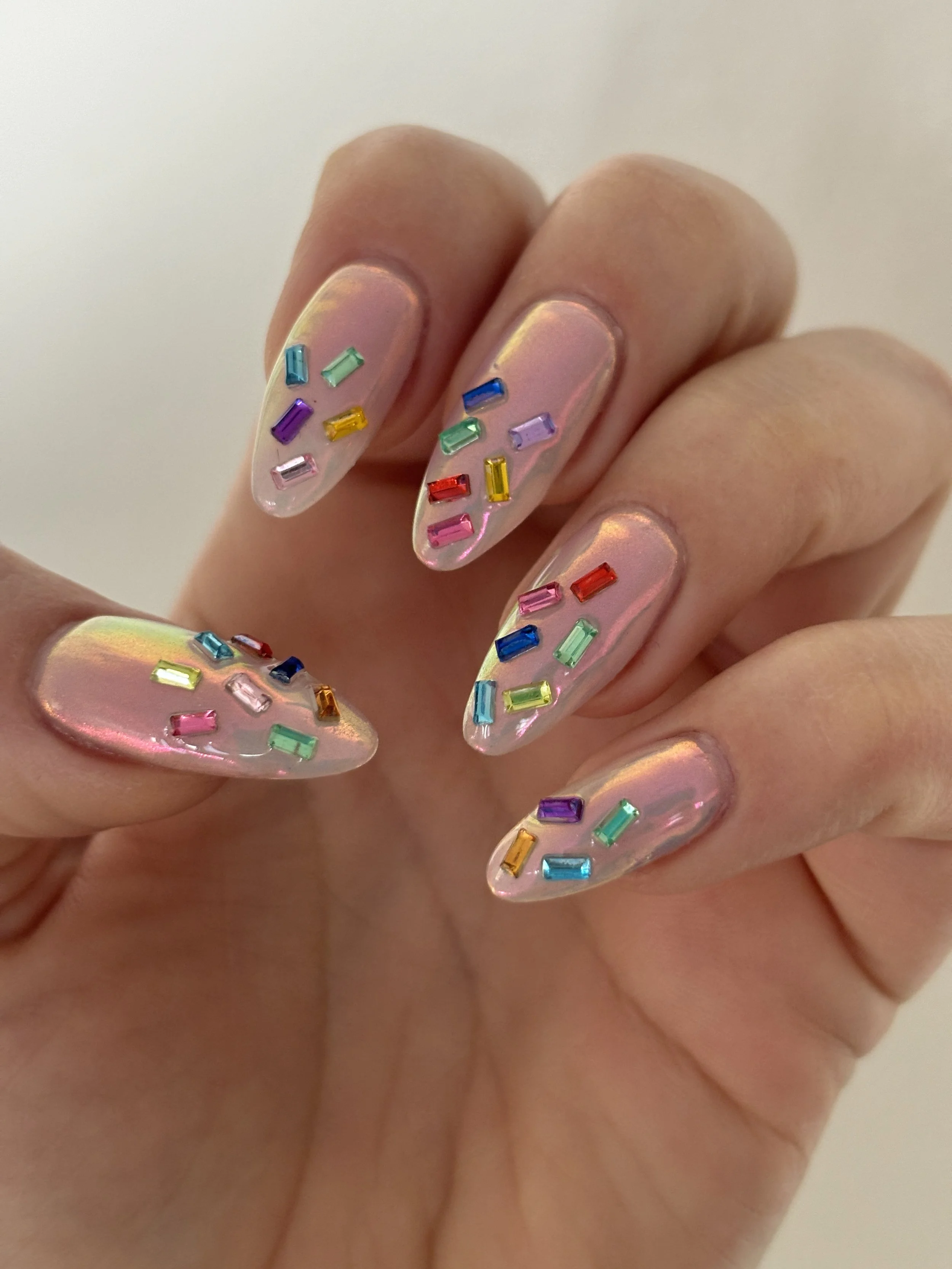 Close-up of a hand with nails painted in a holographic style, decorated with small, rectangular rainbow-colored rhinestones.