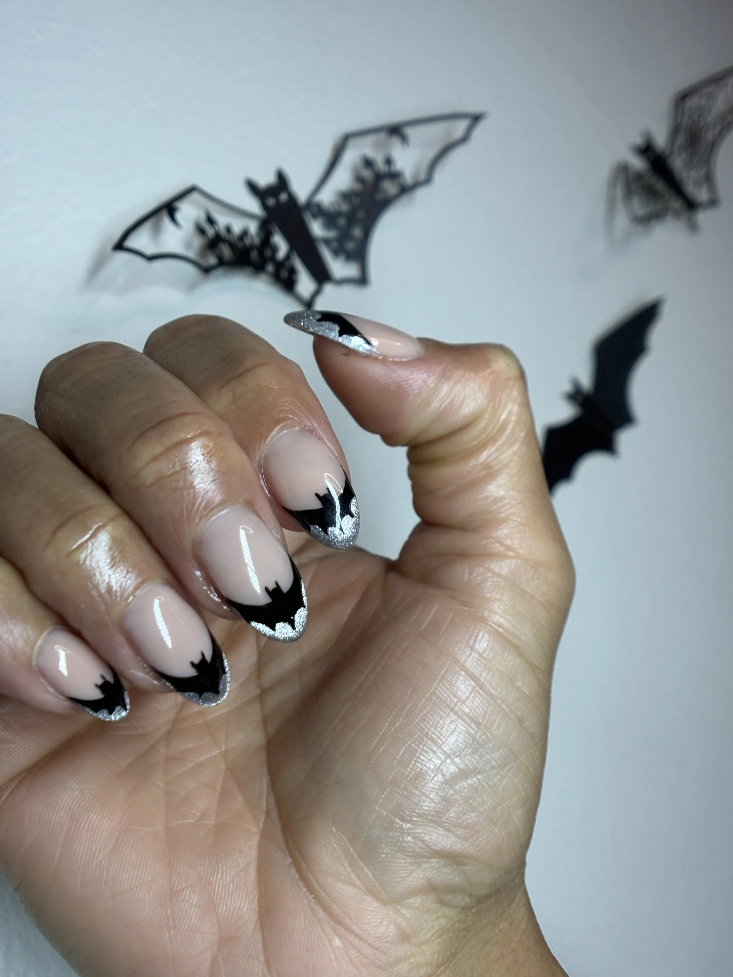 Close-up of a hand with Halloween-themed nail art, featuring black, white, and silver colors with bat and star designs. The person is holding small bat-shaped paper cutouts with a light background.