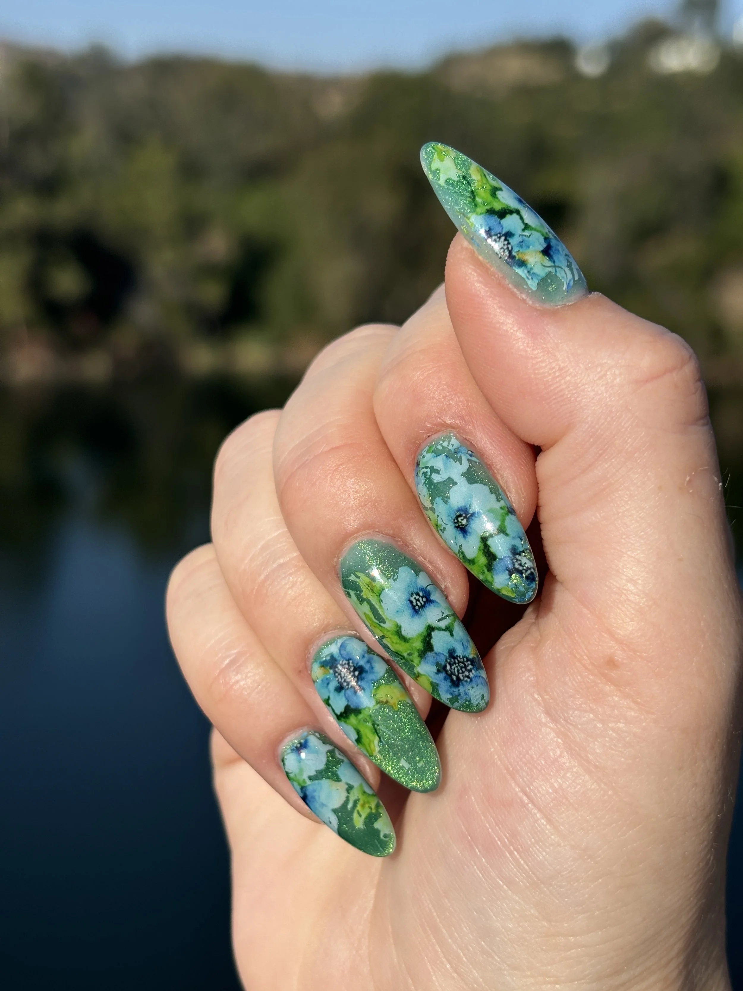Close-up of a hand with long, almond-shaped nails painted with a marble and glitter design in shades of blue, green, and white, held in front of a blurred natural background.
