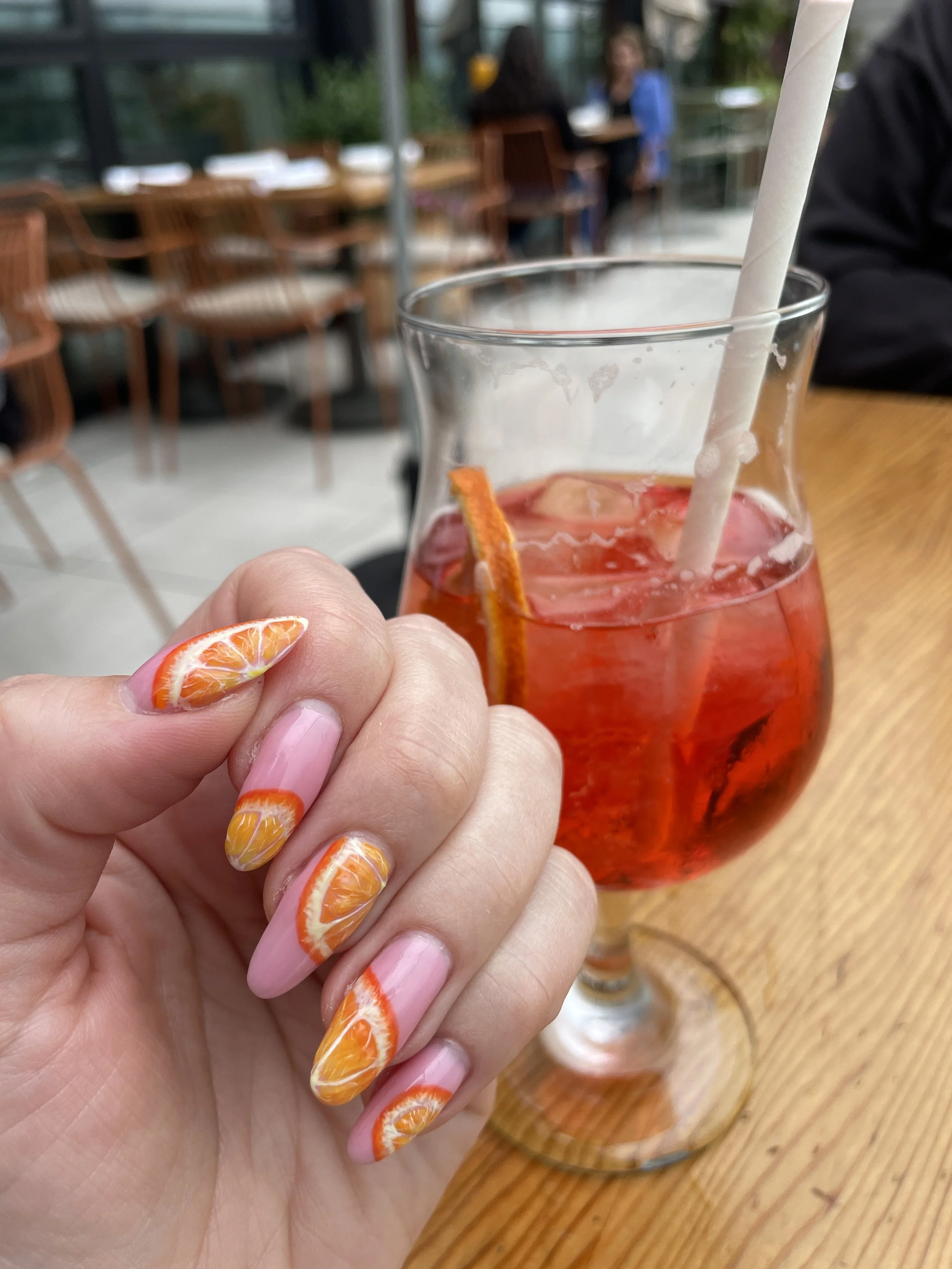 Close-up of a hand holding a glass of pink beverage with a straw and a orange slice garnish. The background shows a blurred indoor dining area with wooden chairs and tables.
